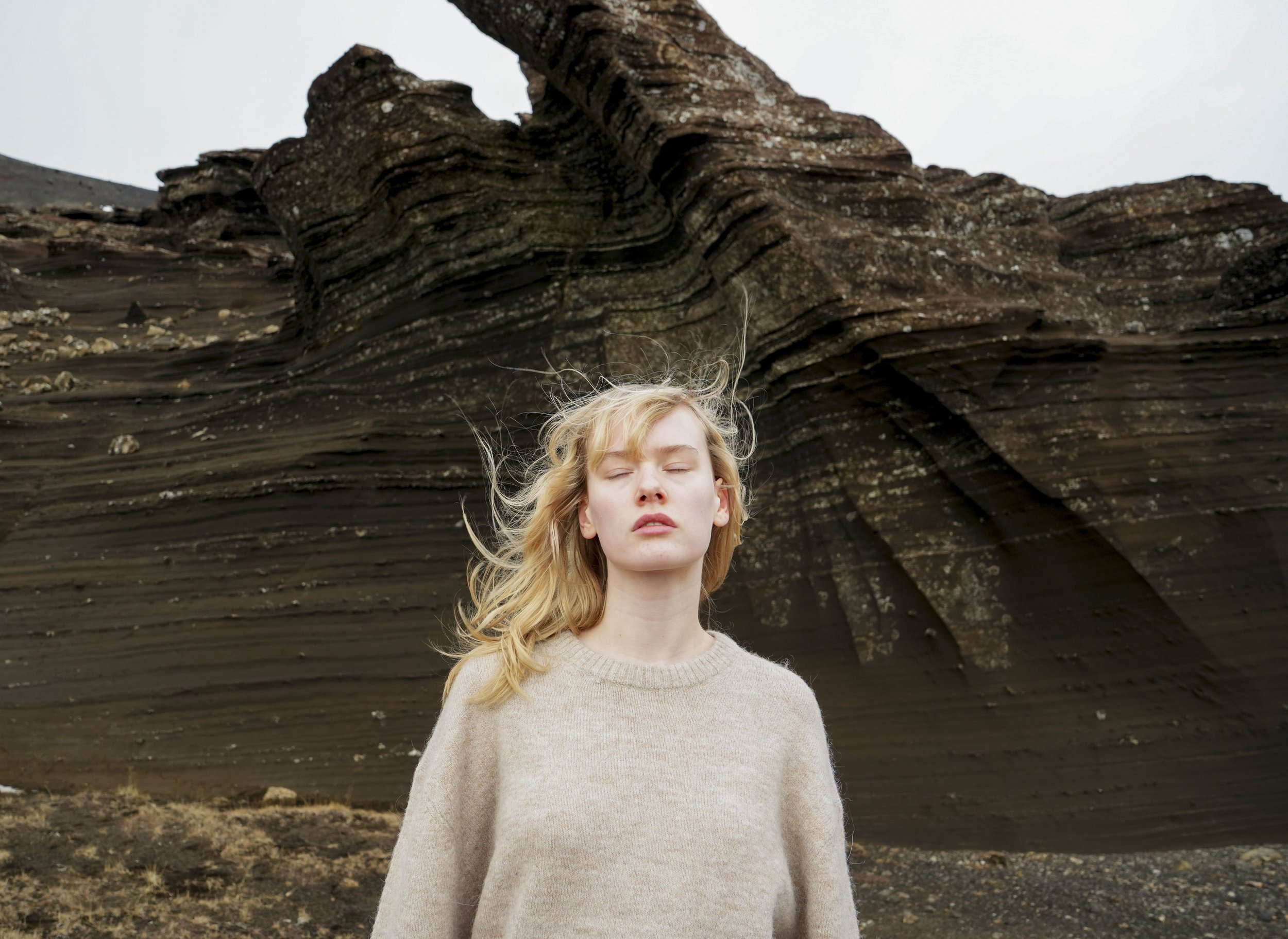 A young woman with blonde hair and closed eyes standing in front of a large, layered rock formation in a barren landscape.