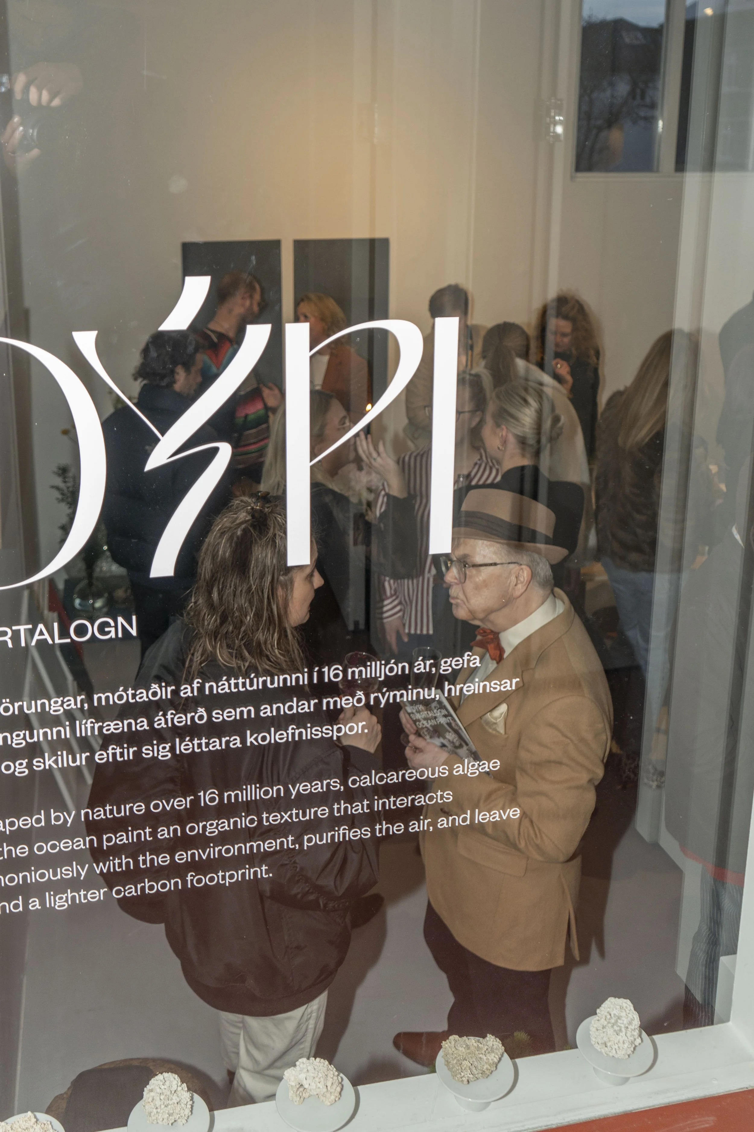 People gathered inside a room, viewed through a glass window with text, at an event or exhibition.