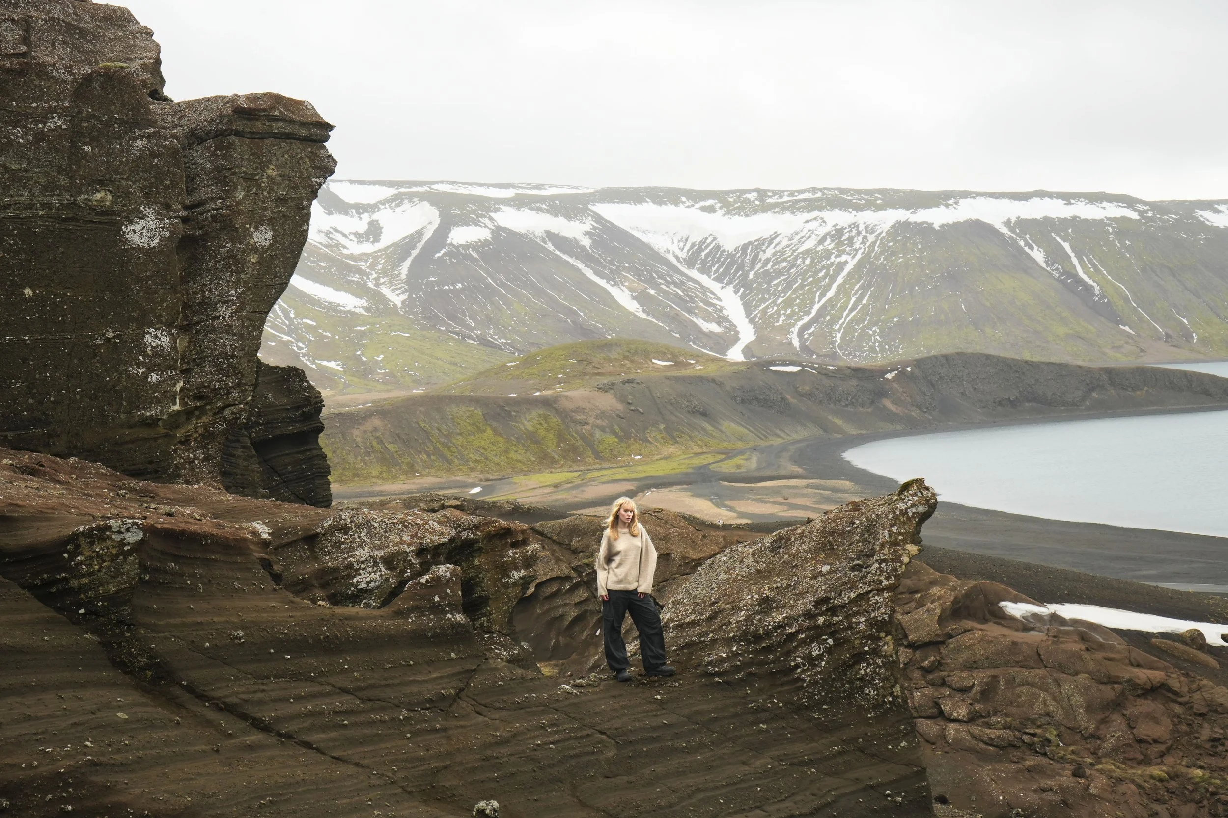 A person standing on rocky terrain with a mountainous, snow-capped landscape and lake in the background.