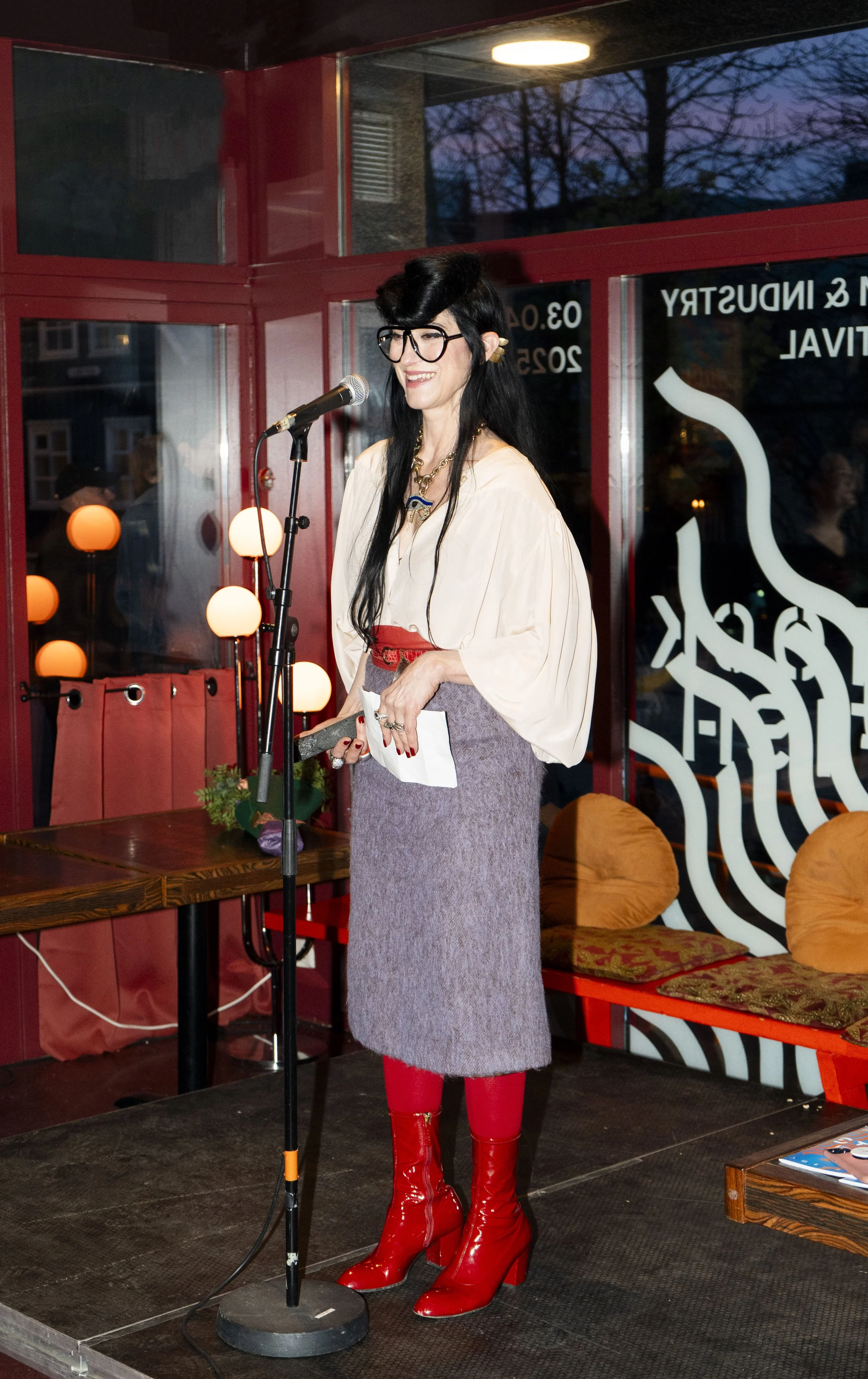 A woman, Floria Sigismondi, with long black hair, wearing glasses, a cream blouse, a gray skirt, red tights, and shiny red high-heeled boots, standing on a stage with a microphone in front of her, smiling. 
