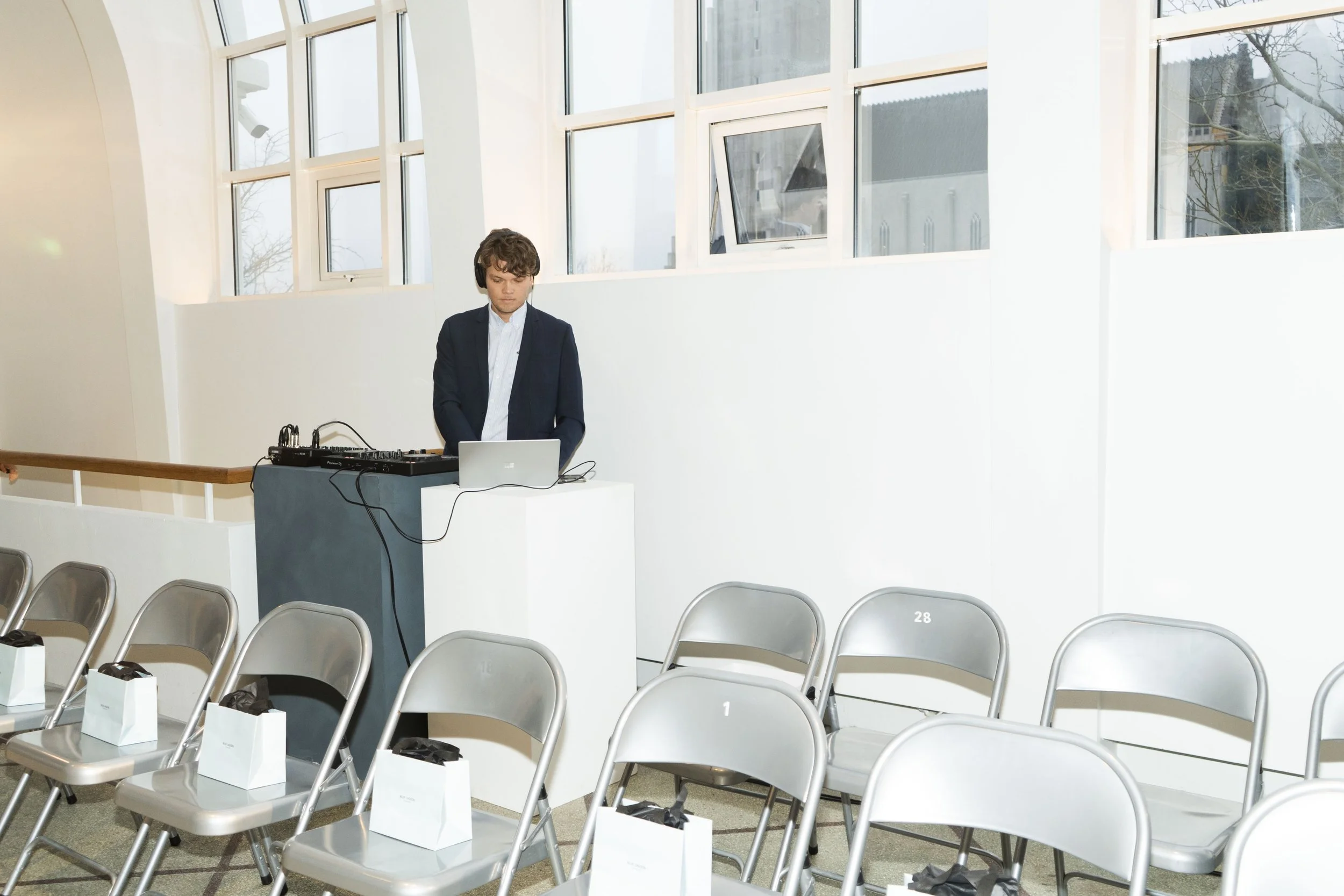 A young man in a suit stands behind a DJ setup with a laptop and equipment in an empty event hall with white walls and large windows. Rows of empty chairs with white gift bags are in the foreground.