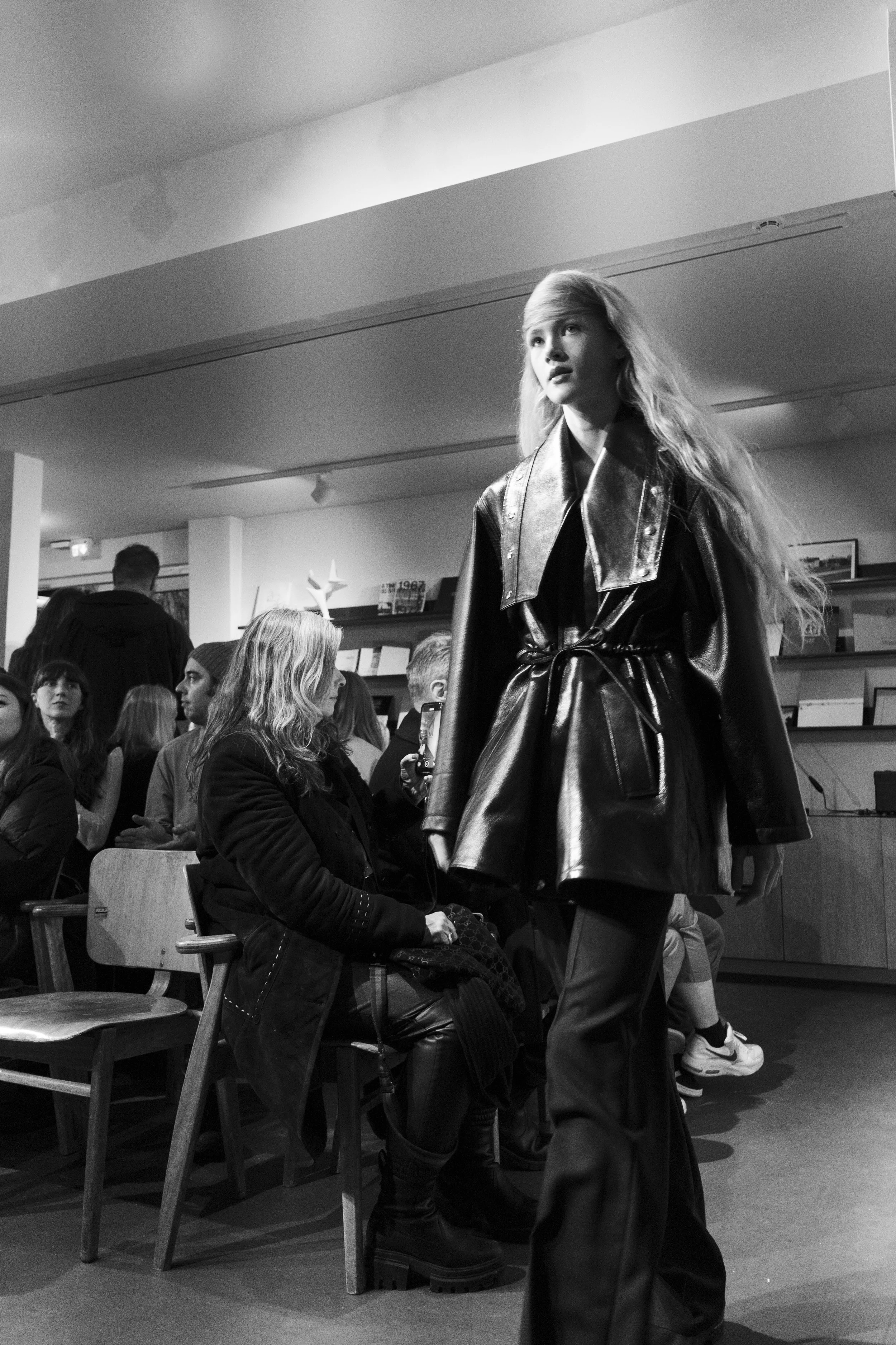 A young woman walking in a crowded indoor space, wearing a shiny black leather coat and loose pants, with long wavy hair, in black and white photography.