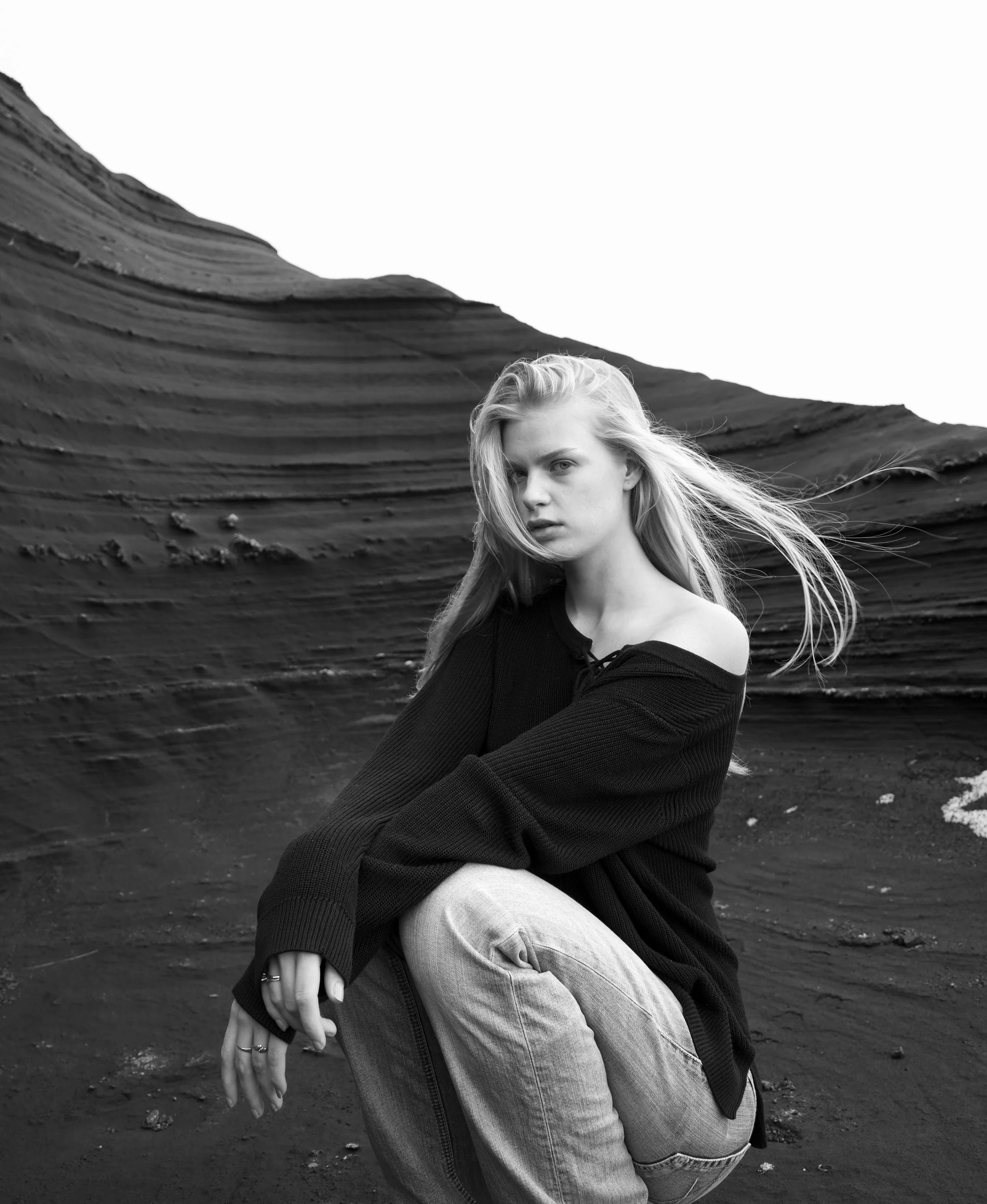 A young woman with long blonde hair squatting on dark, rocky terrain with layered rock formations in the background, wearing a black off-the-shoulder sweater and light-colored jeans, looking at the camera