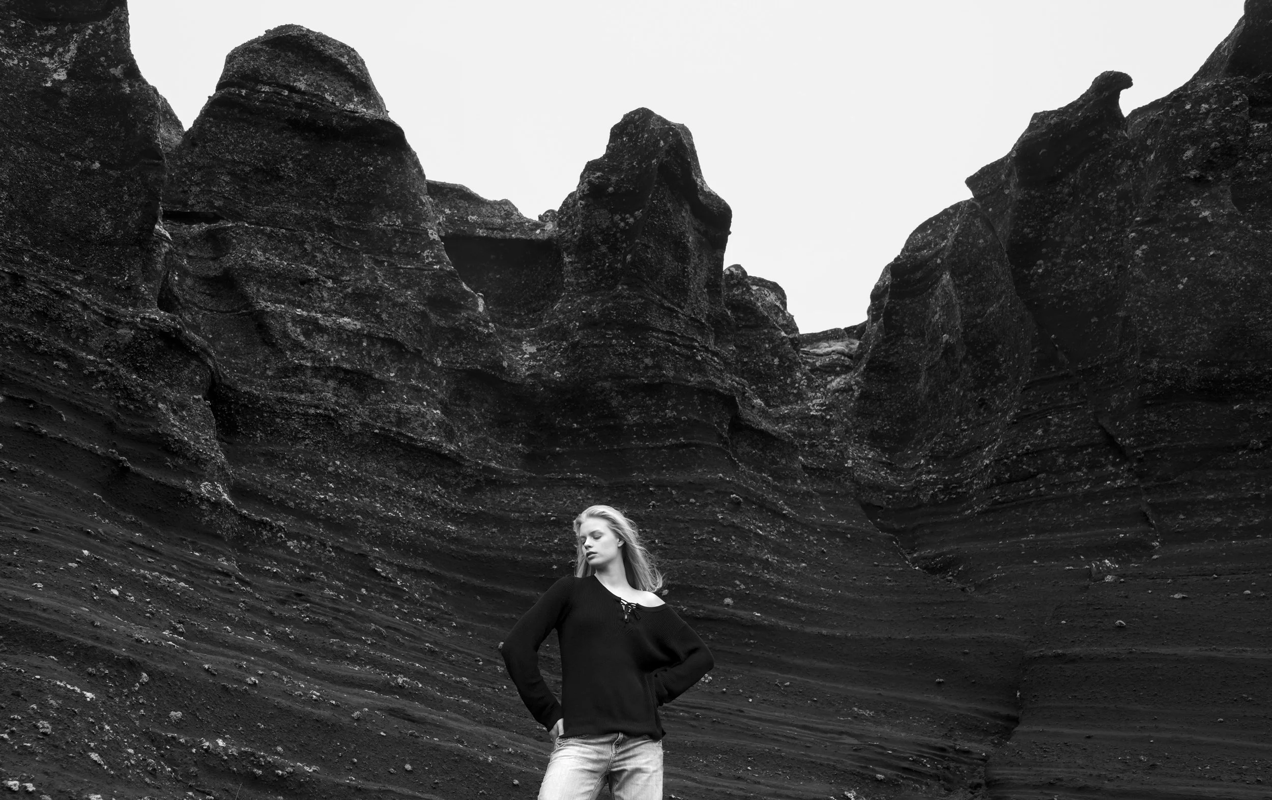 Black and white photo of a woman standing near large, layered rock formations with towering peaks in the background.