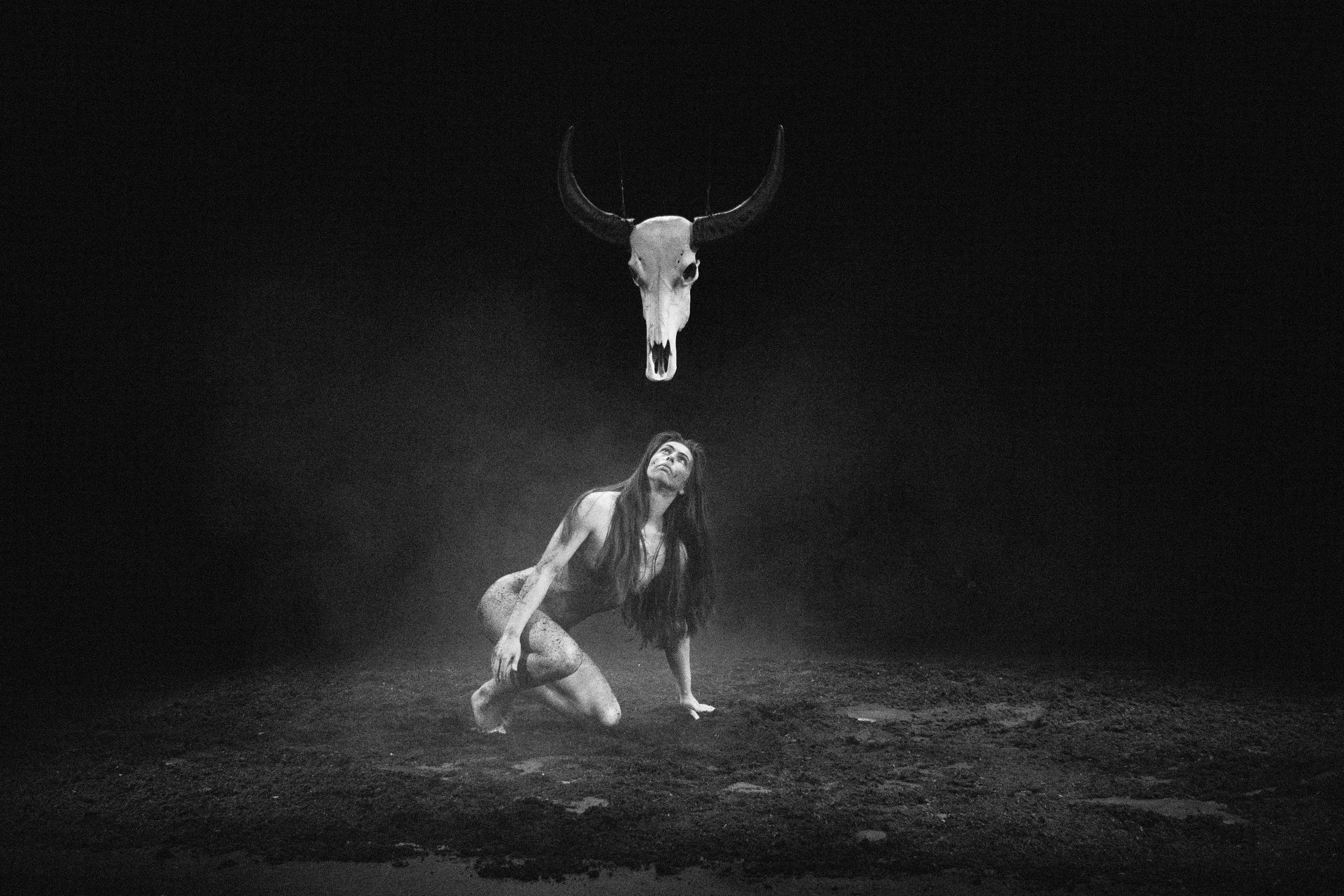 A black and white image of a woman with long hair, kneeling on the ground, looking up at a large animal skull with horns hanging from the ceiling.