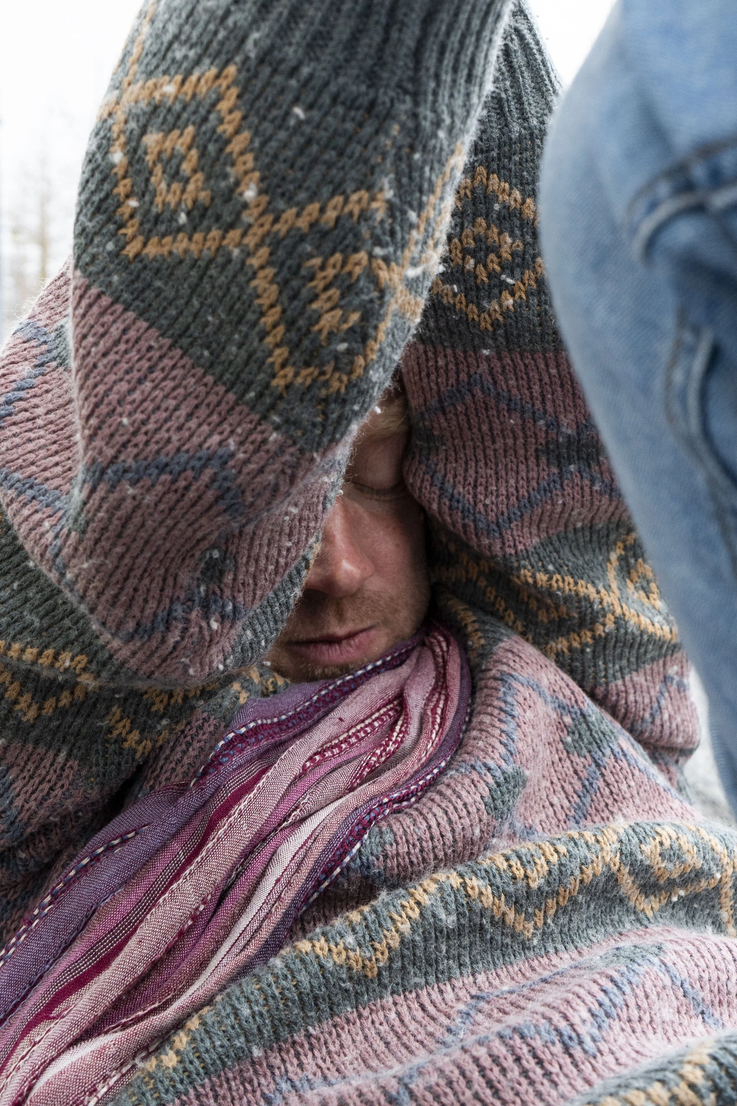 Person with light skin and blond hair, wearing a patterned sweater, with their head bowed and eyes closed, outdoors in cold weather.