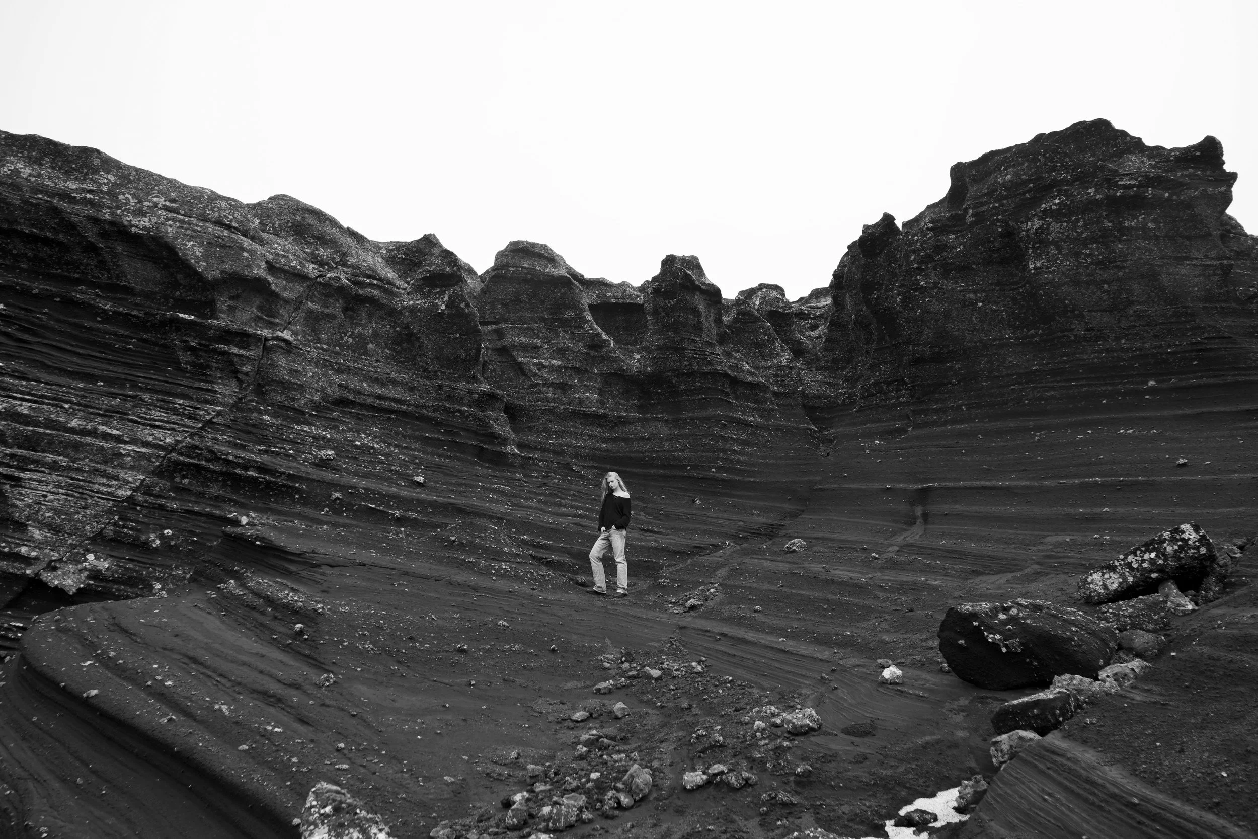 A person standing in a black and white rocky canyon with layered, textured cliffs and a cloudy sky above.