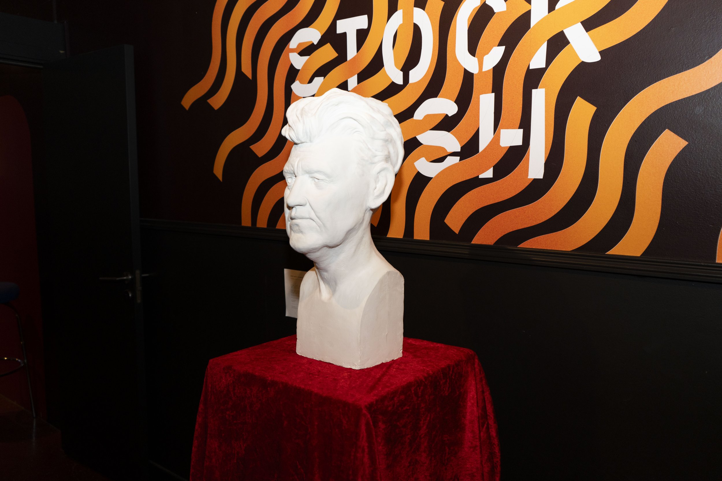White bust sculpture of David Lynch with styled hair placed on a red velvet-covered pedestal, against a dark wall with orange and white wavy line design