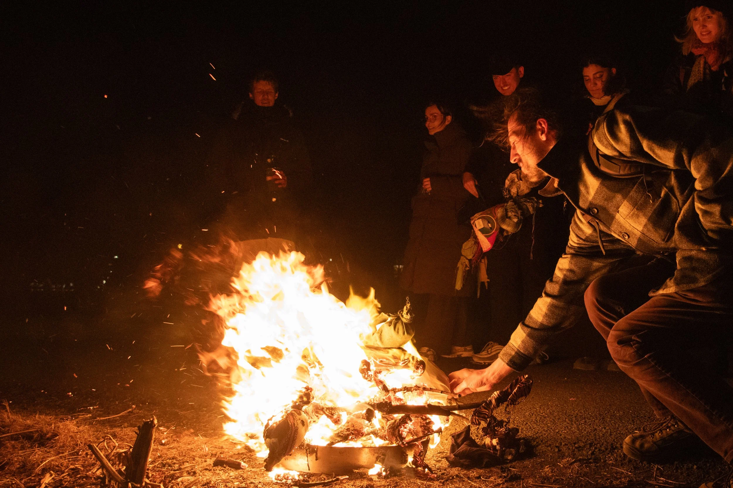 Group of people gathered around a campfire at night, with one person tending the fire. Marzanna. Morana