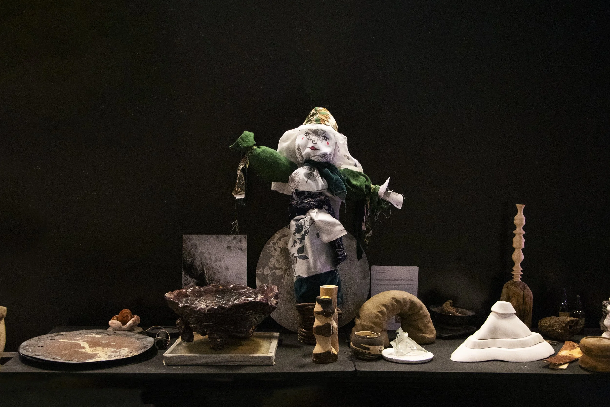 Artistic display with fabric doll, Marzanna, pottery, sculpture, and various objects on a black table against black background.