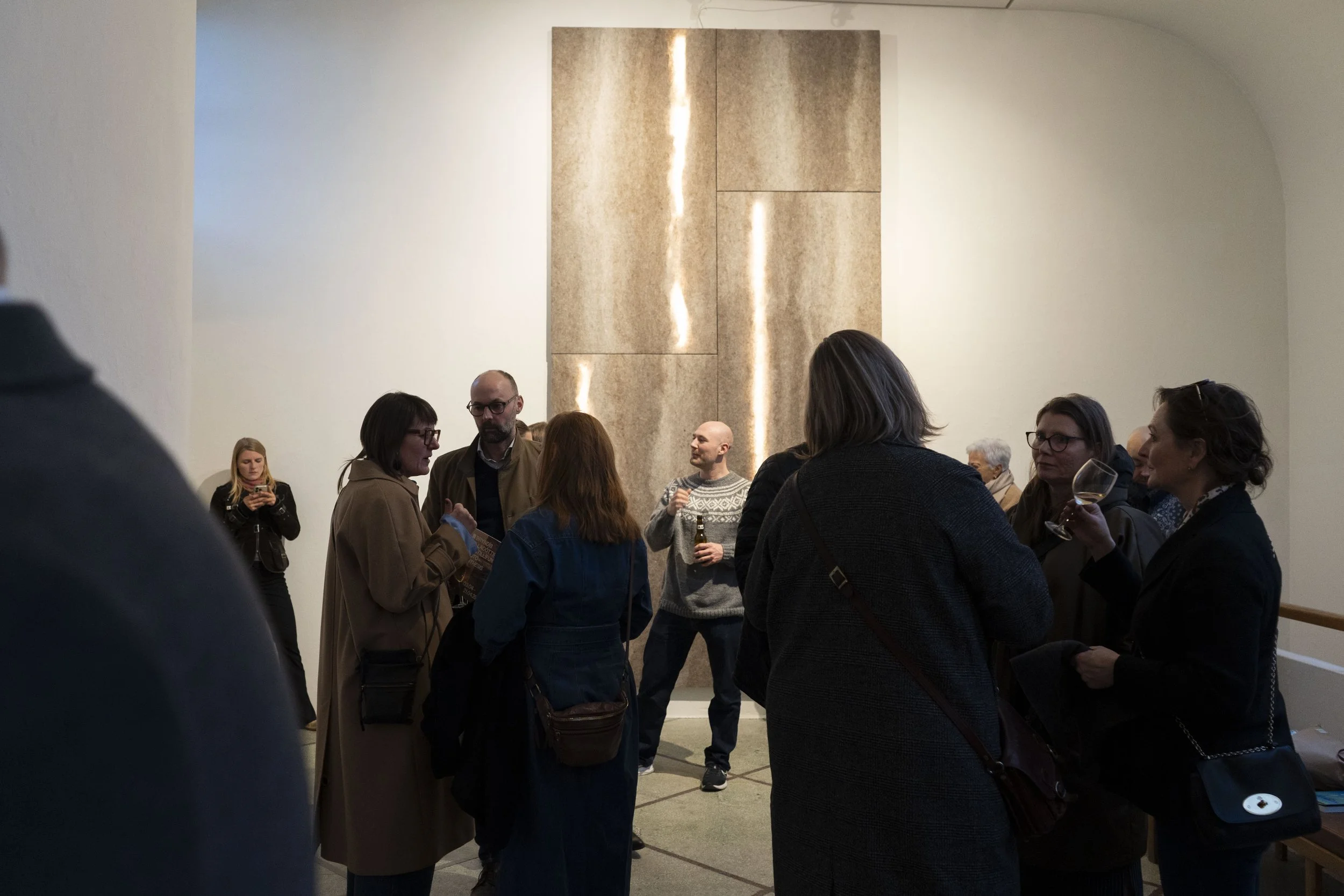 Group of people socializing at an indoor event, standing and holding drinks, with a large artistic wall behind them.