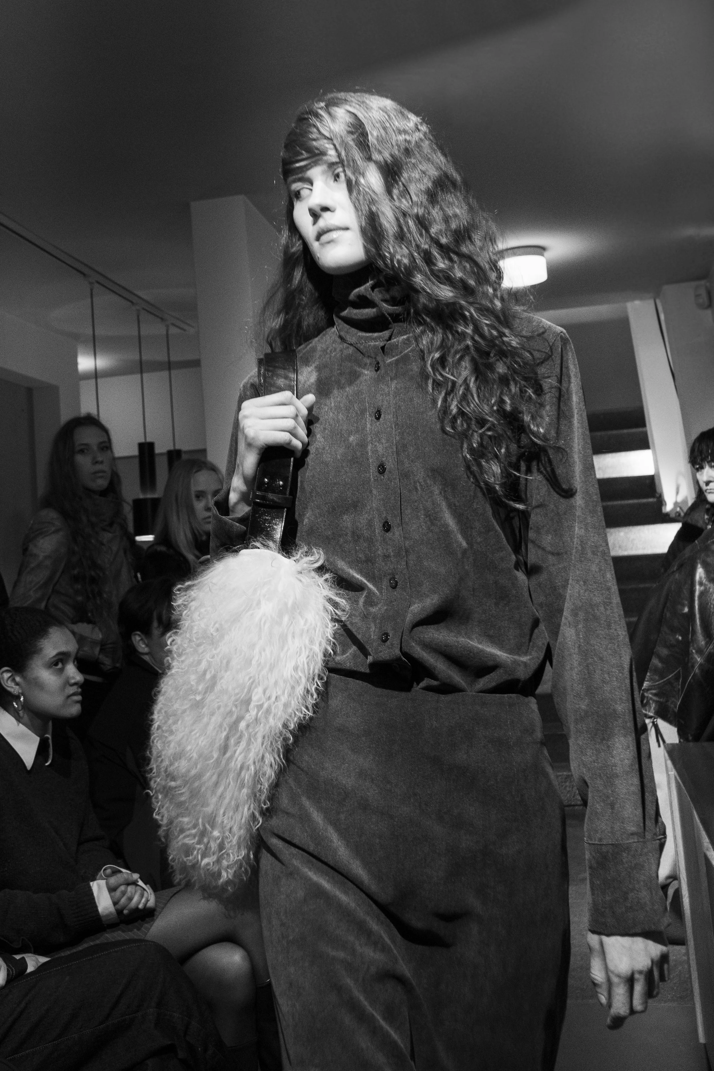 Black and white photo of a young woman with long curly hair walking in a fashion show, wearing a dark button-up shirt and high-waisted belt, carrying a fluffy white bag over her shoulder, with an audience of young women seated and watching in the bac