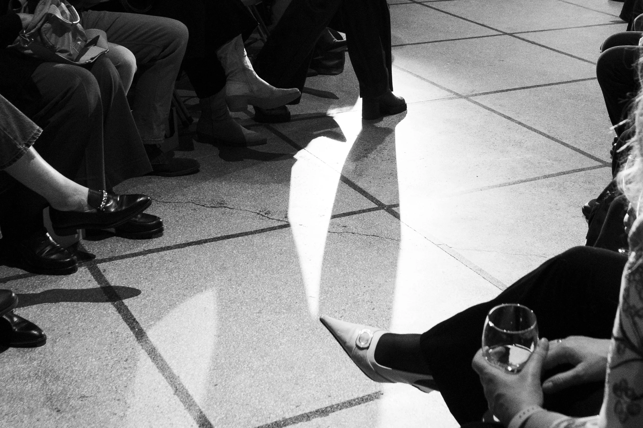 People sitting in a circle on chairs, with a reflection of their legs and a woman holding a glass of wine seen on the tiled floor in black and white.