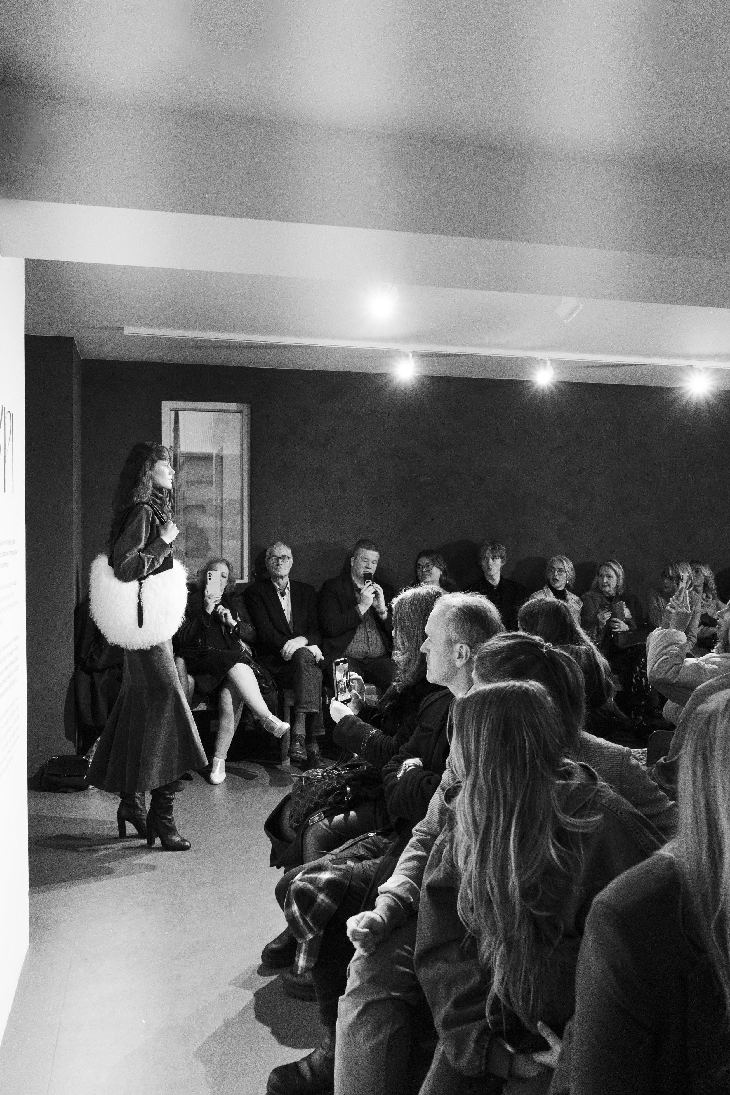 A woman speaking to an audience in a gallery or exhibition space, with many in the audience taking photos or listening attentively.