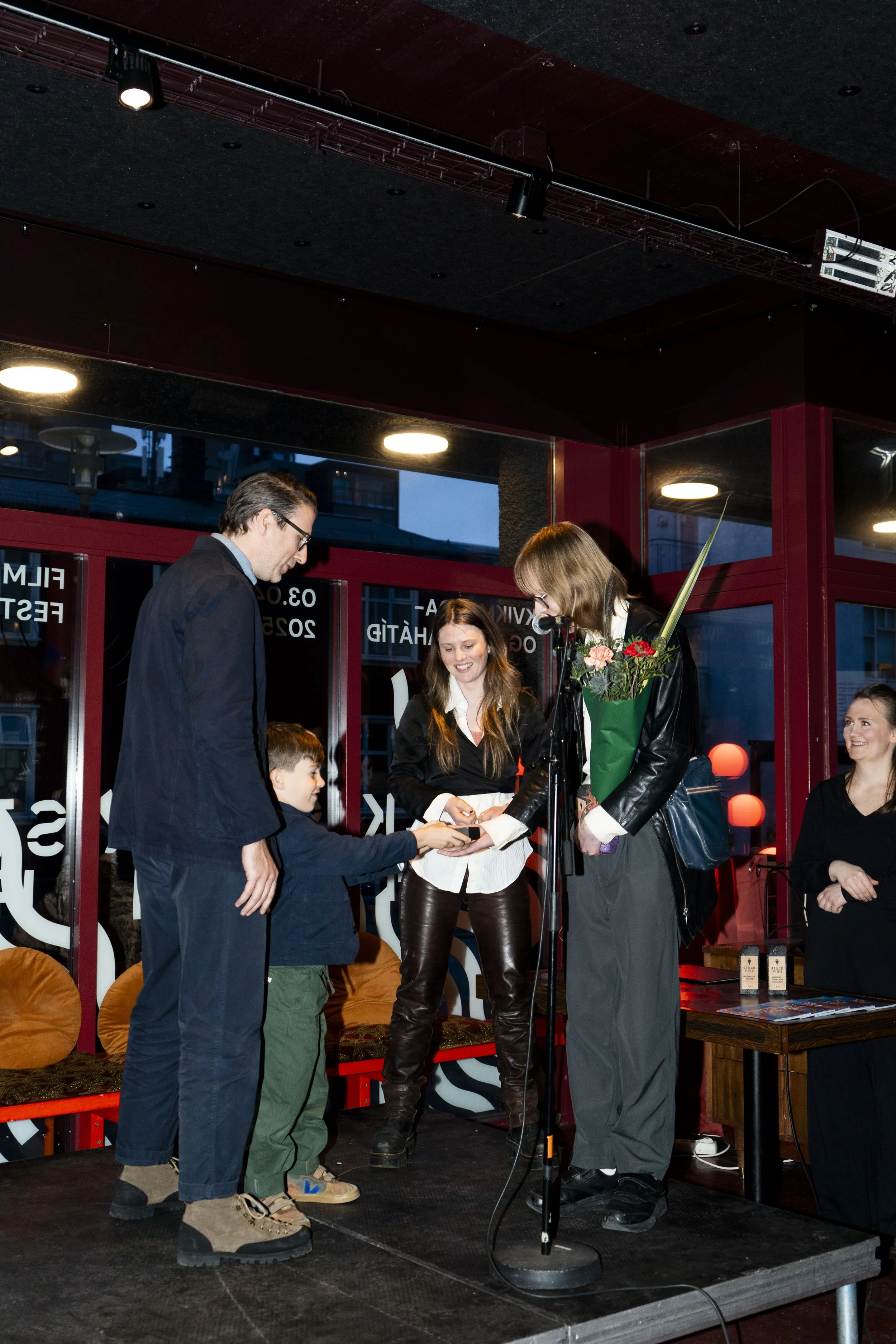 A woman receiving an award or recognition on stage from a man, with a woman holding flowers standing next to her. Two children are also present, one handing something to the woman. They are in a dimly lit indoor setting, possibly a restaurant or even