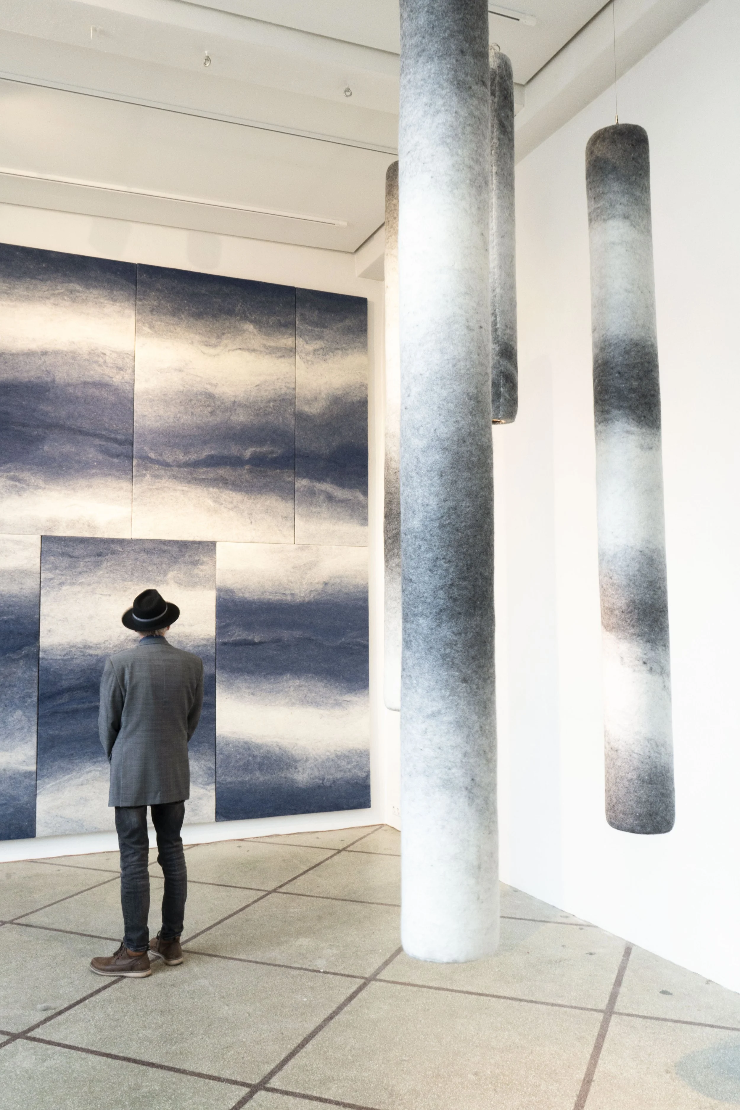 A person wearing a gray blazer, jeans, and a hat stands in an art gallery looking at a large abstract blue and white painting on the wall, with three gray cylindrical sculptures hanging from the ceiling nearby.