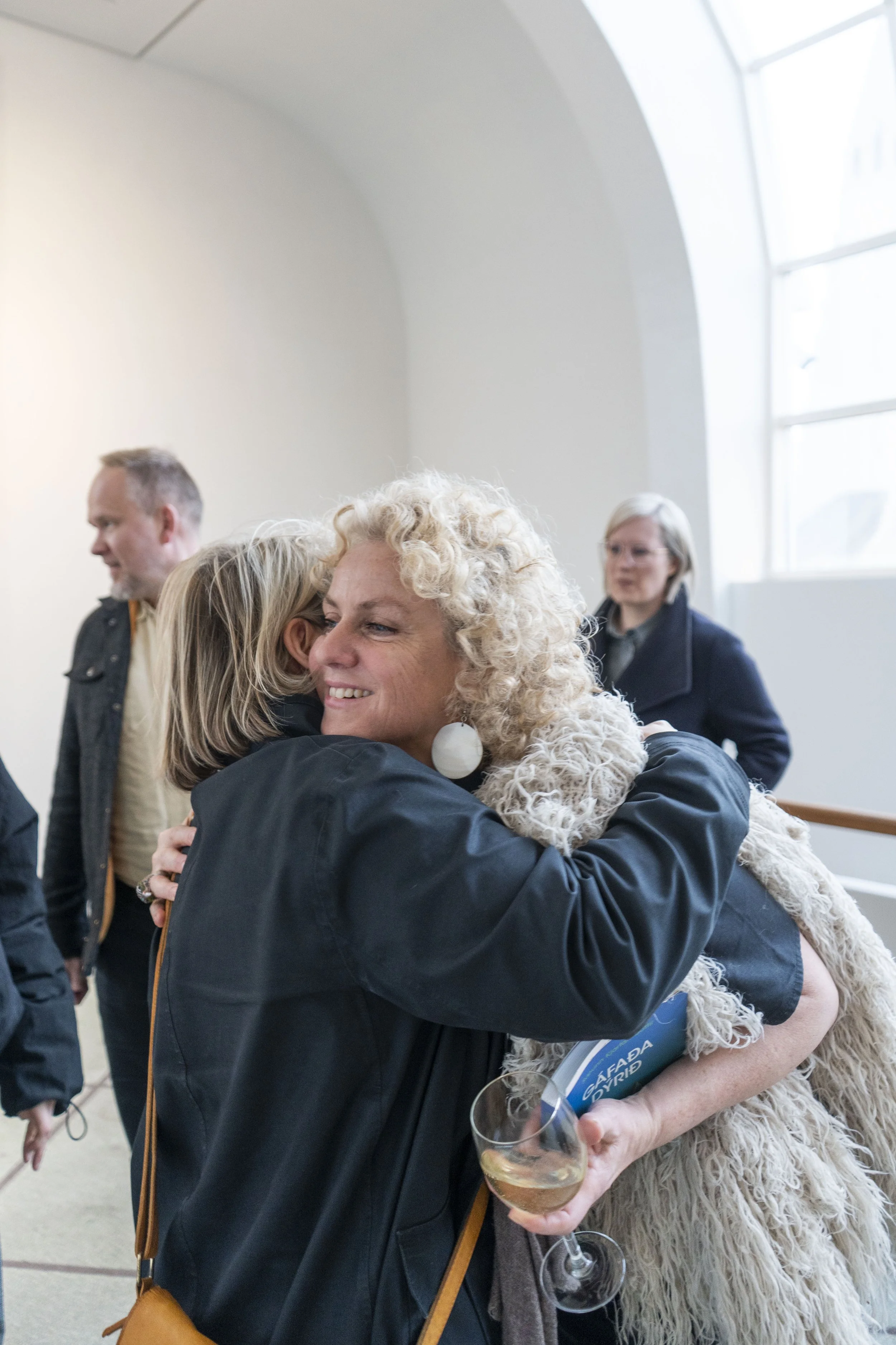 Two women hugging, one holding a glass of wine, at a social gathering indoors, with other people in the background.