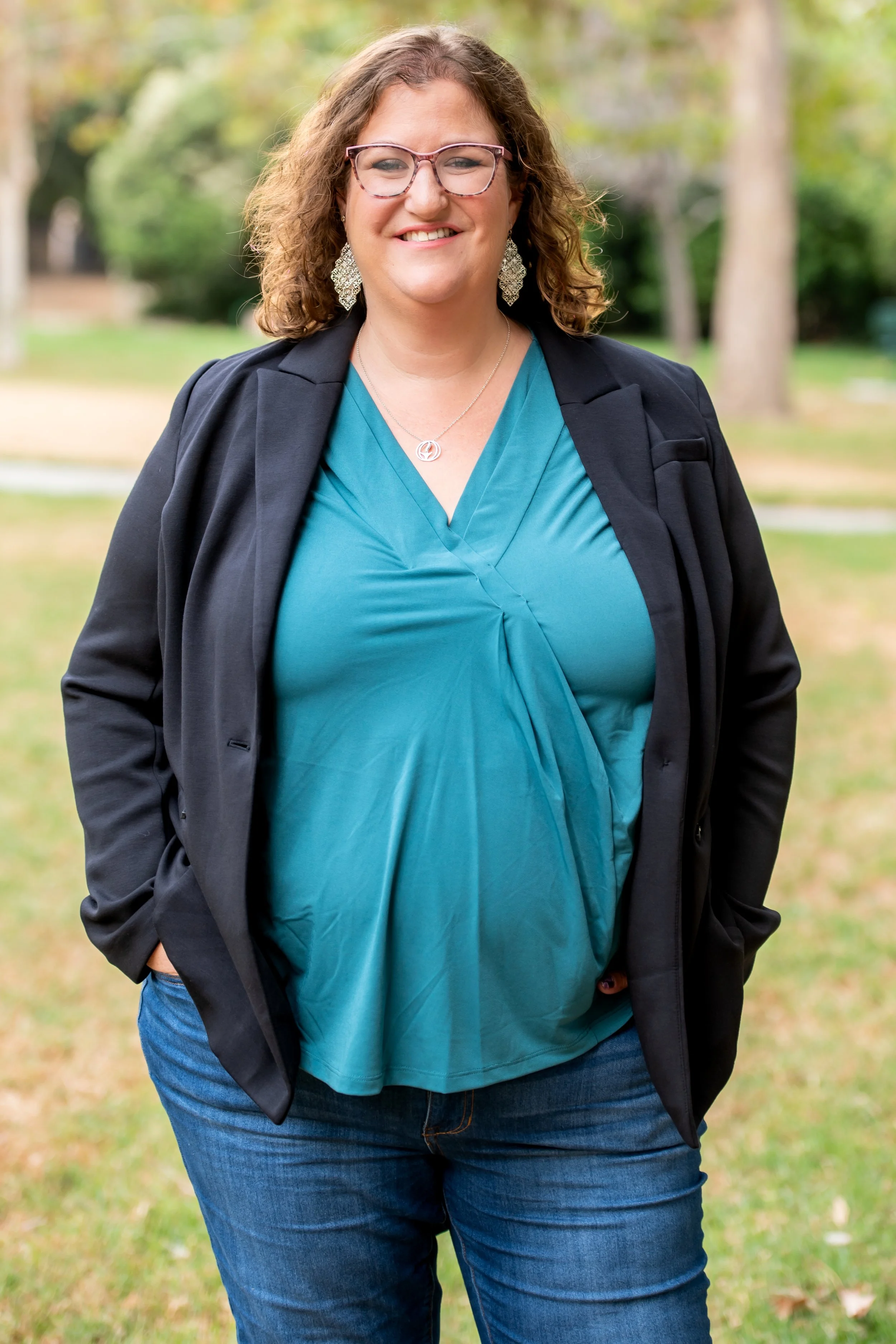 White woman wearing a teal top and black jacket. She has curly hair and glasses and is smiling