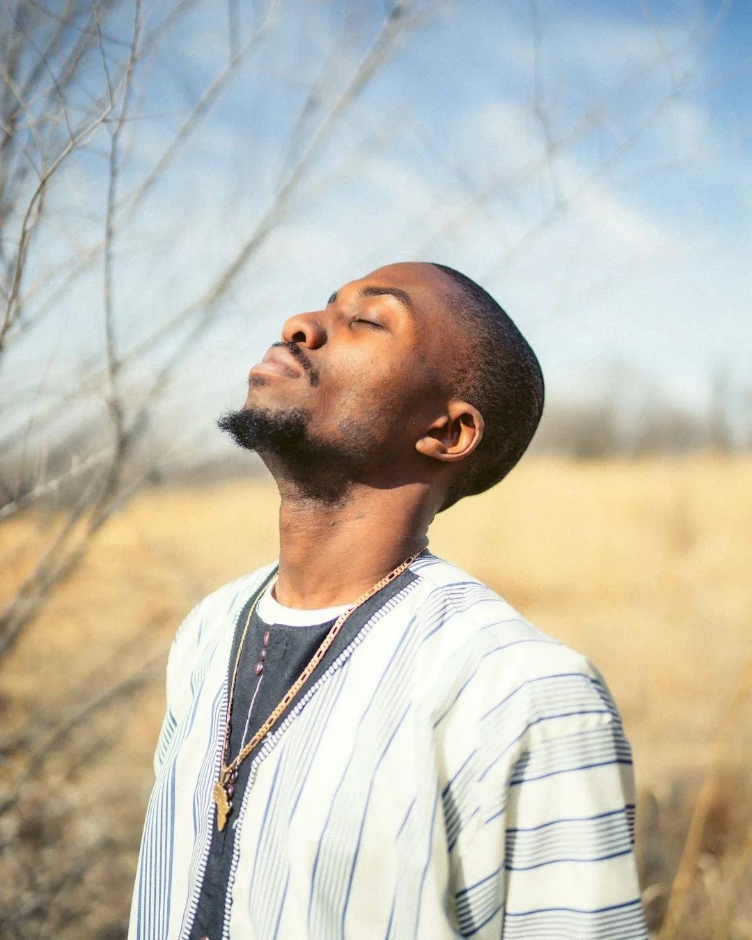 A man with closed eyes and relaxed expression, standing outdoors in a field with dry grass and bare trees, enjoying sunlight on his face.