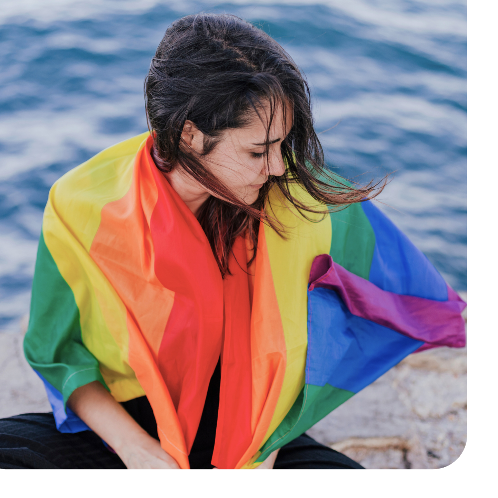 Woman with dark hair wrapped in a rainbow pride flag, sitting by the water.