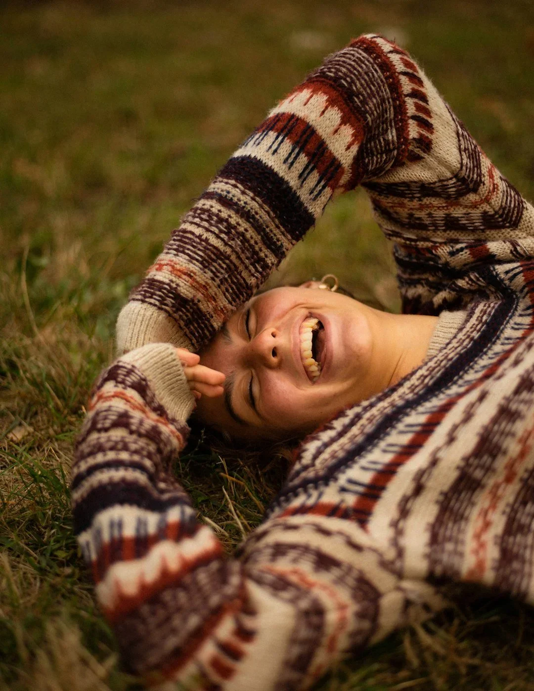 A person wearing a patterned sweater lying on grass, laughing with eyes closed and hand on forehead.