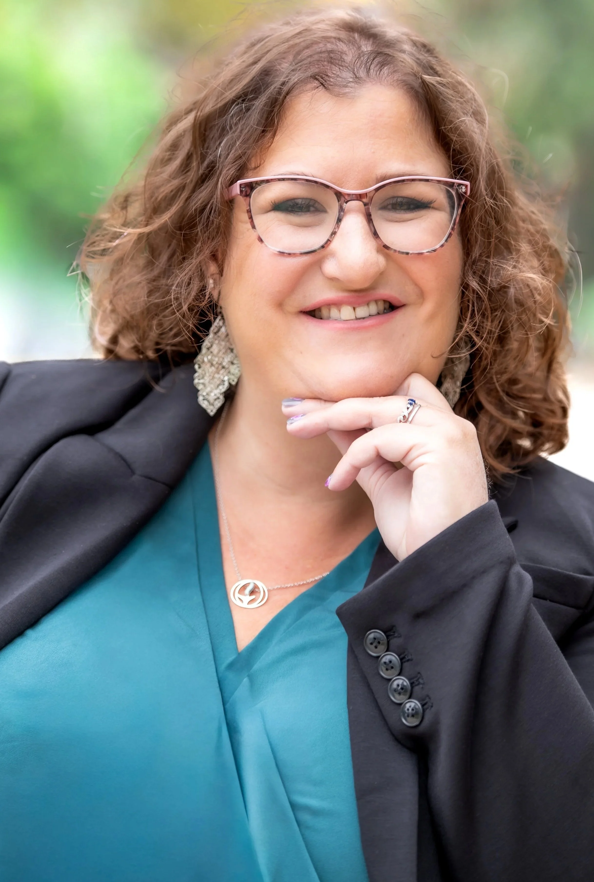 A woman with curly brown hair and glasses, smiling outdoors. She wears a teal top, a black blazer, statement earrings, and a necklace with a peace sign pendant. She rests her chin on her hand.