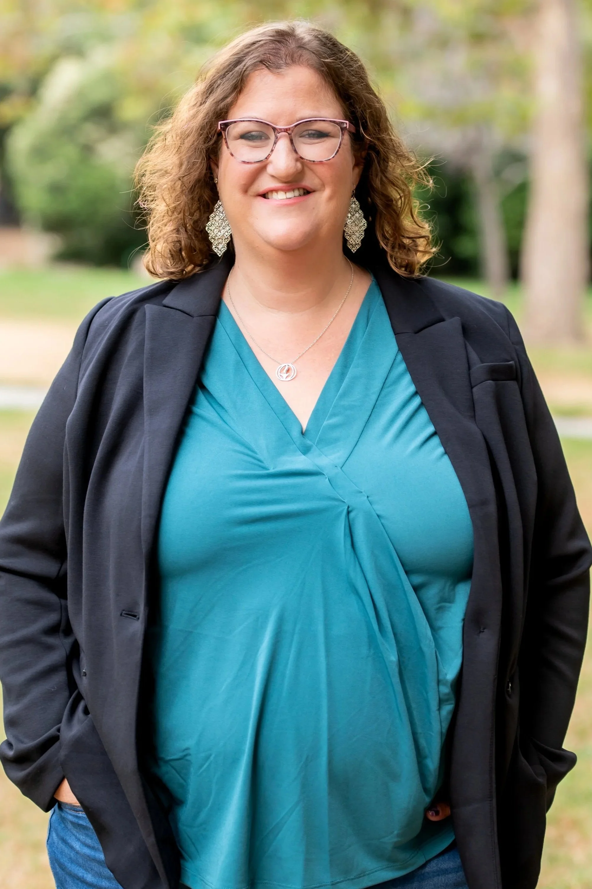 White woman wearing a teal shirt and black jacket. She is smiling and wearing glasses