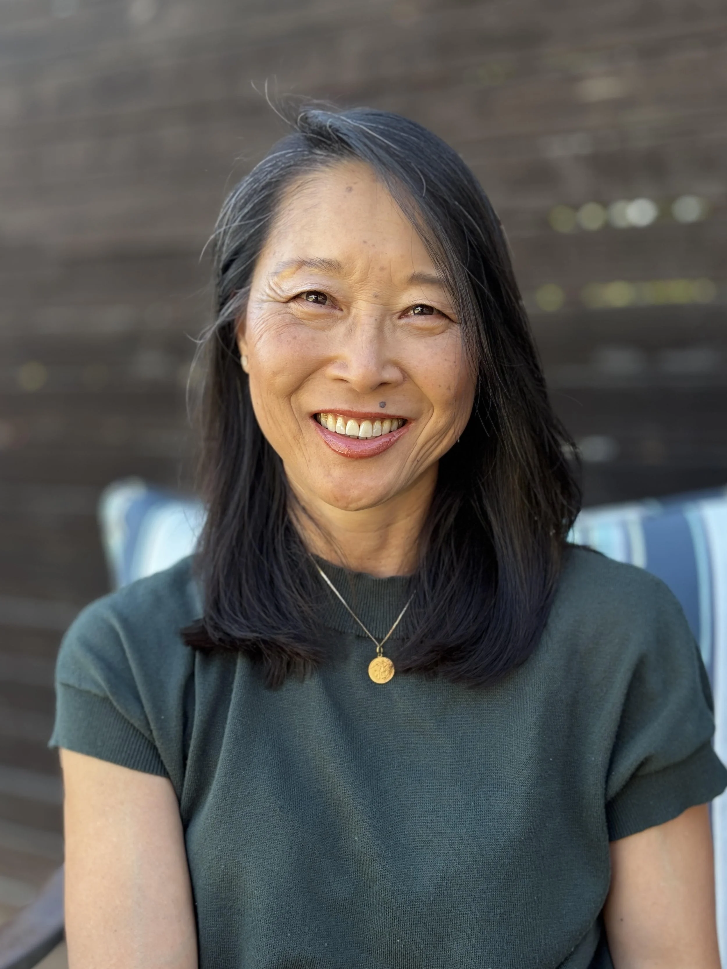 An older Chinese American female wearing a dark teal shirt and a gold necklace. She has black hair that falls just below her shoulders and she is smiling