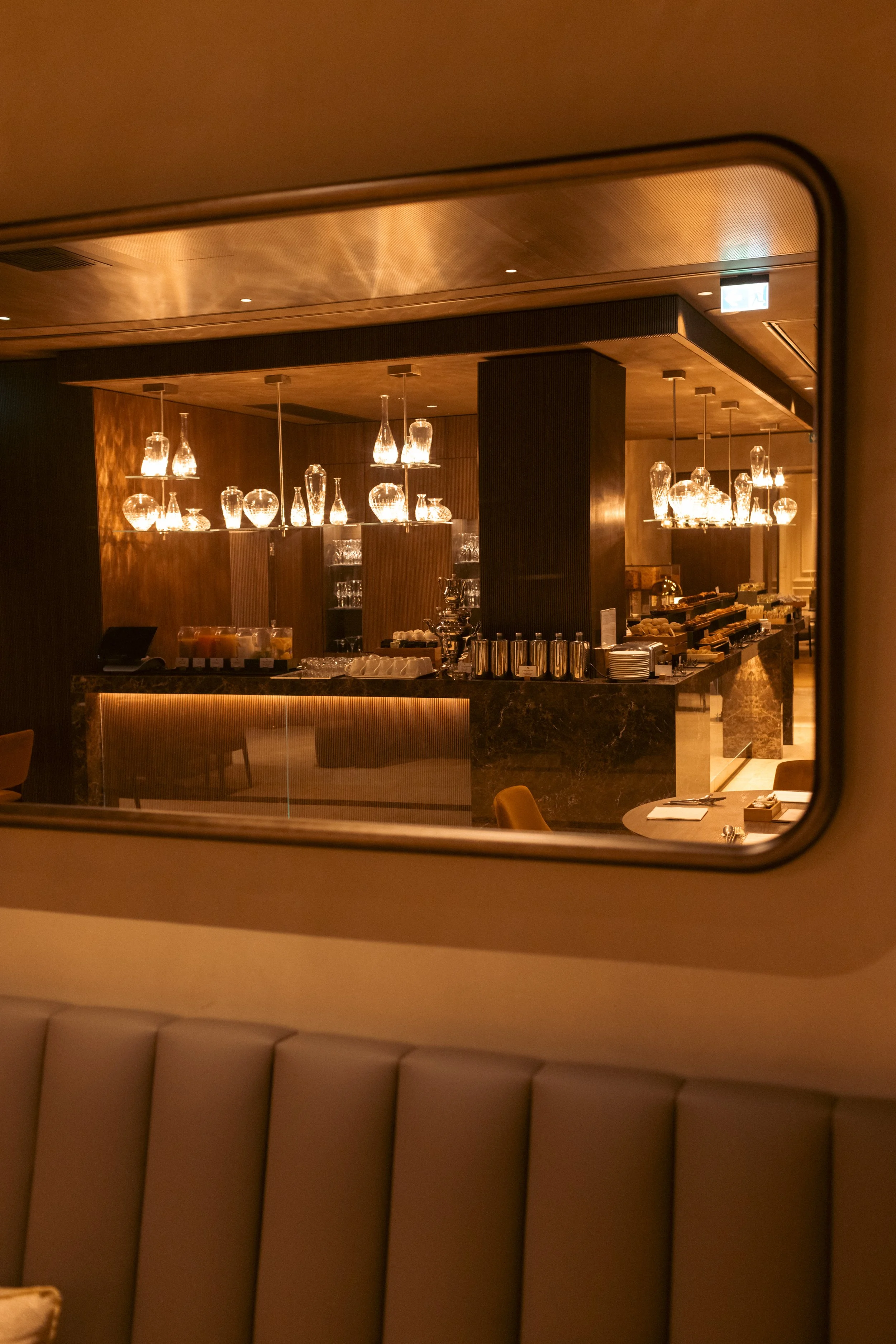 An elegant hotel restaurant with warm lighting, glass vases and bottles on a dark marble counter, wooden panels, and chairs with upholstered backs.
