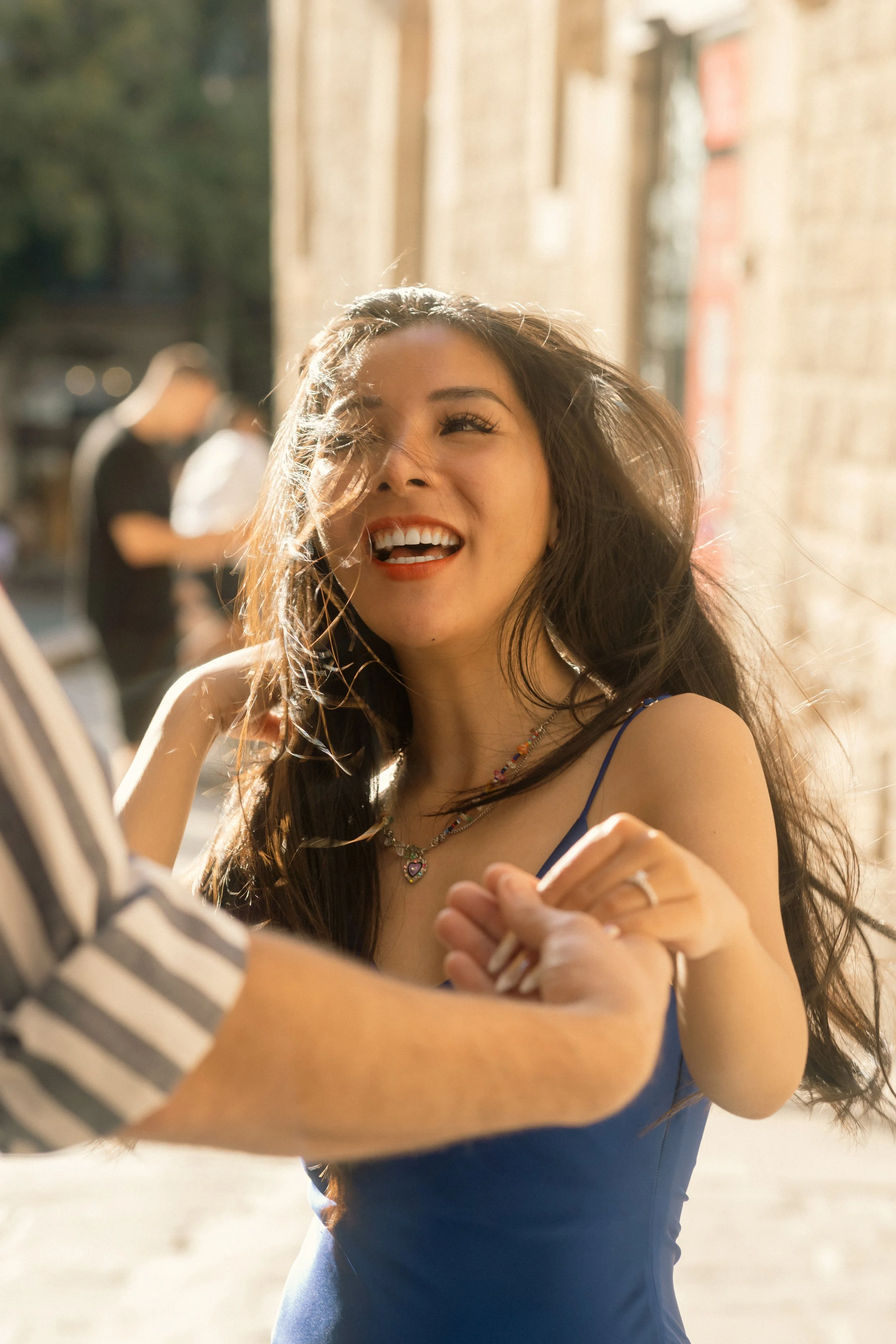 A woman with long dark hair, smiling and laughing, wearing a blue dress and jewelry, dancing outdoors with someone whose arm is visible in the foreground.