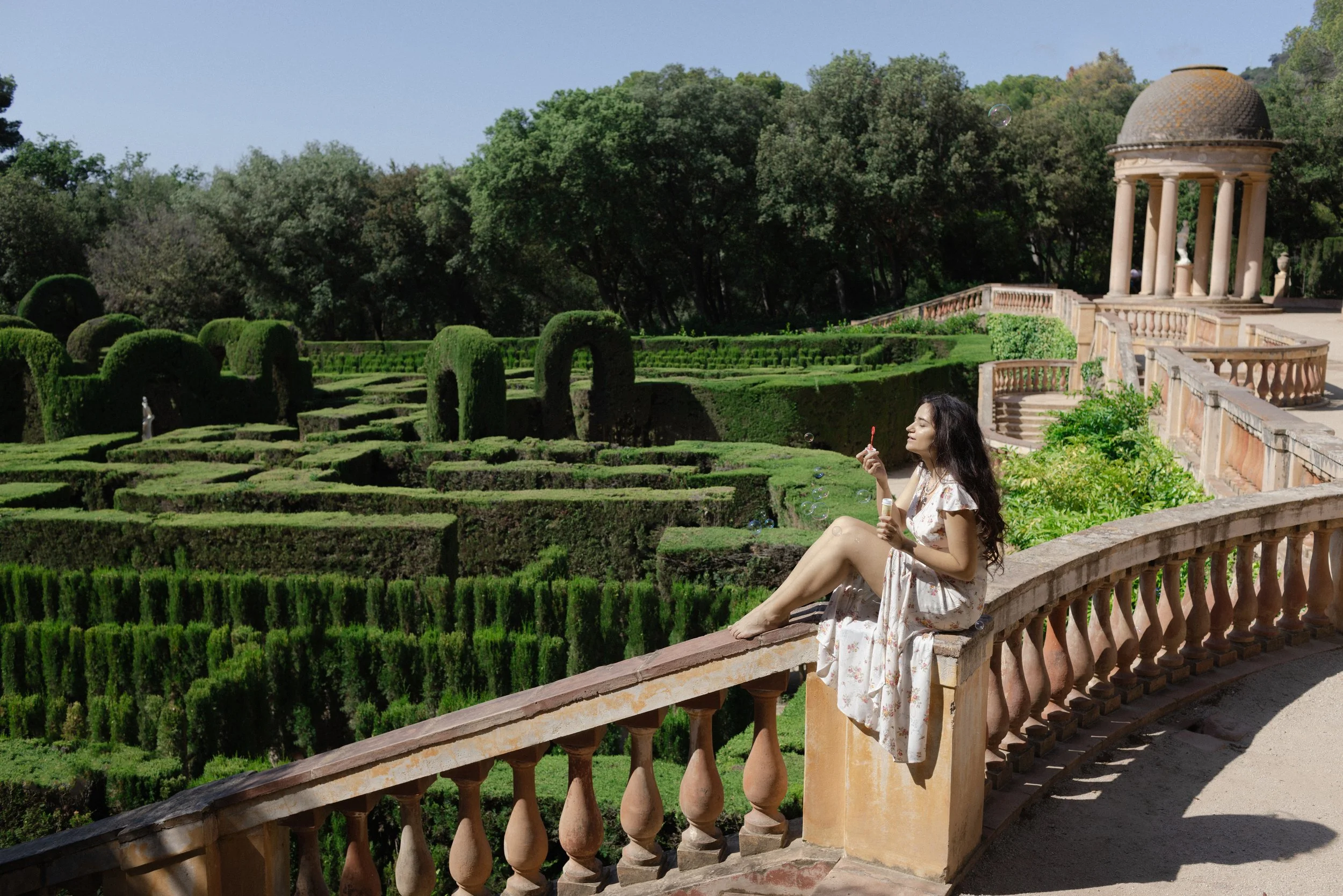 A woman in a floral dress sitting on a stone ledge by the railing, blowing bubbles in a lush, formal garden with manicured hedges and a stone pavilion in the background.