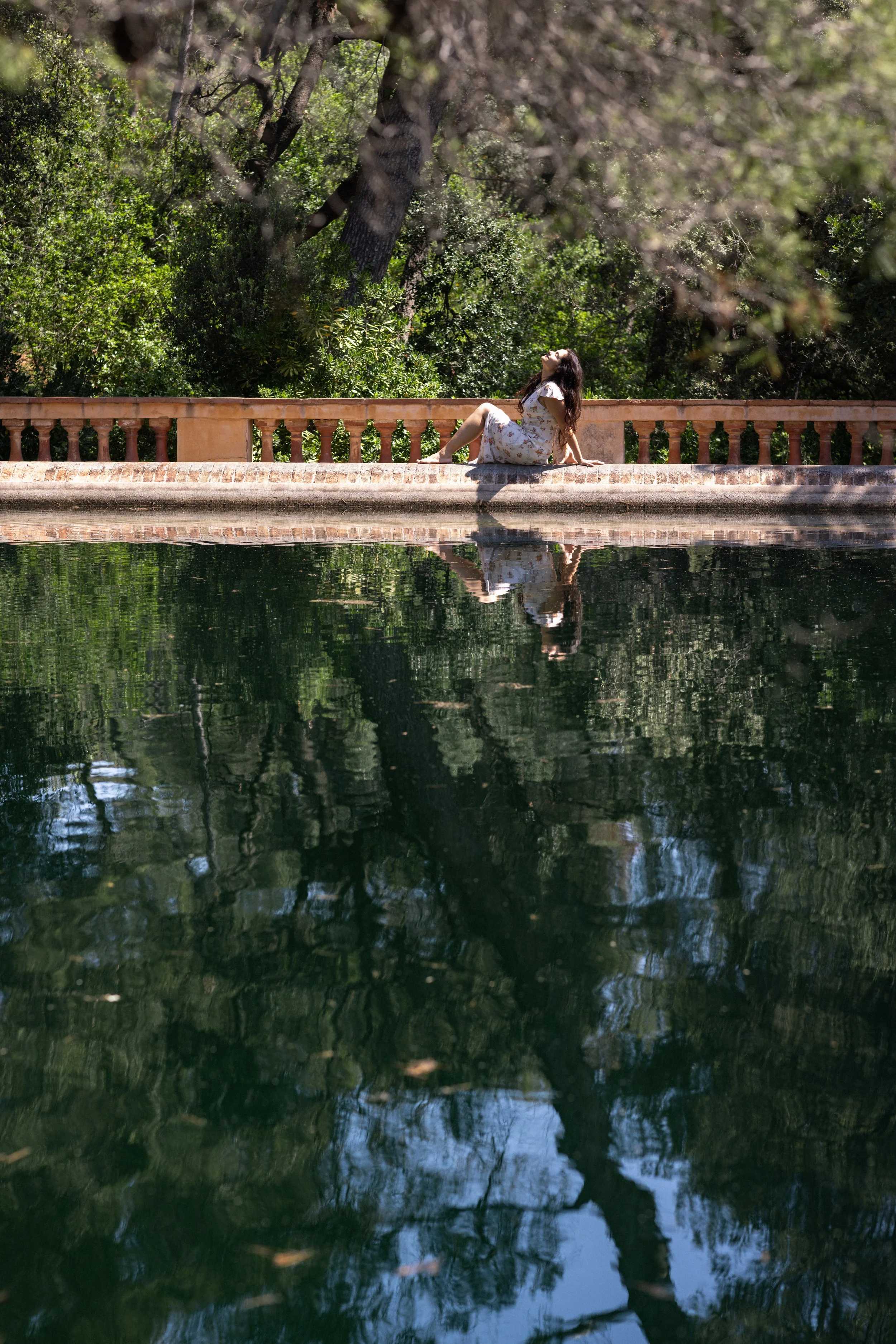 Woman sitting on a brick ledge next to a reflective body of water, with trees and greenery in the background.