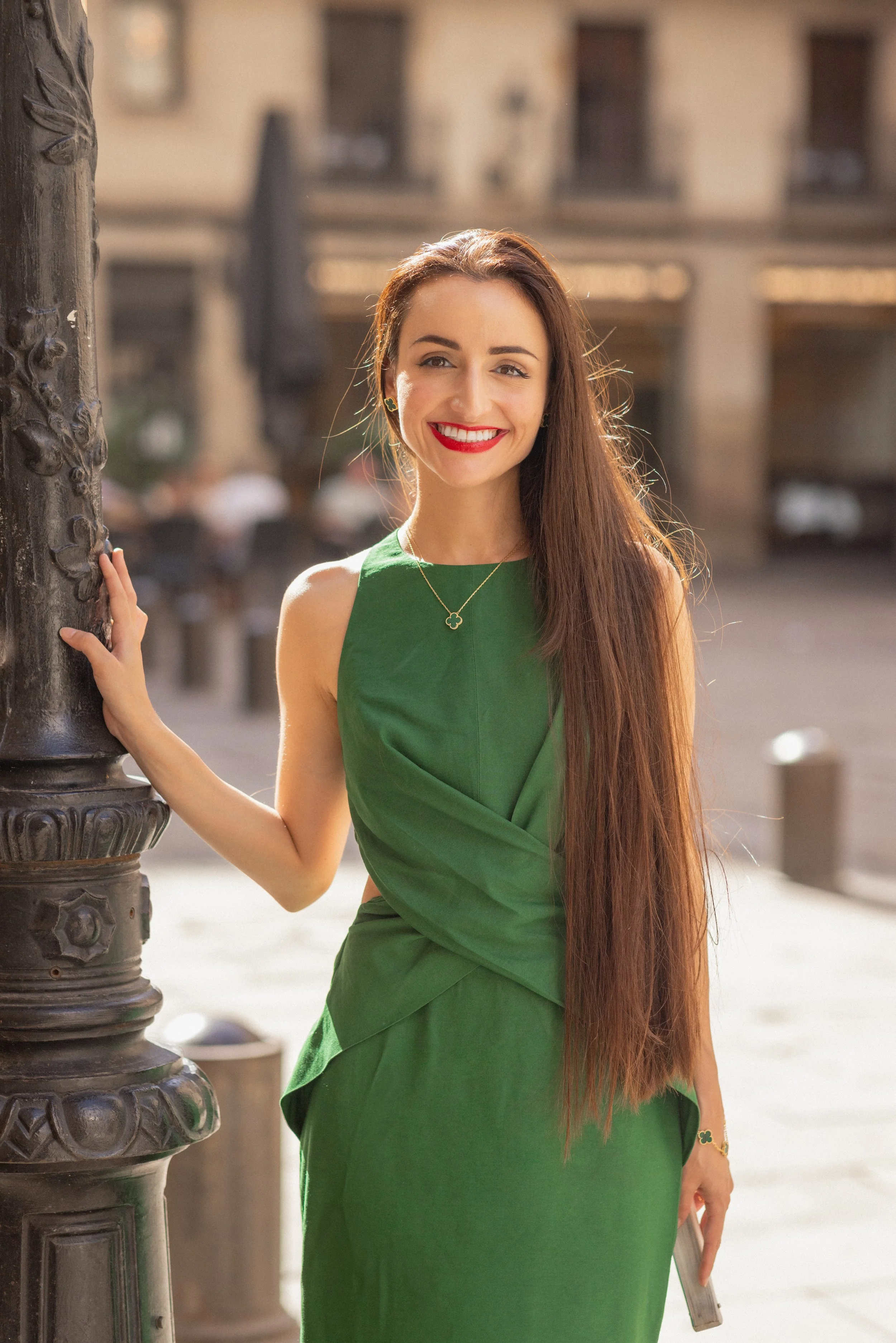 A woman with long brown hair, wearing a green dress, smiling and standing outdoors near a streetlamp.
