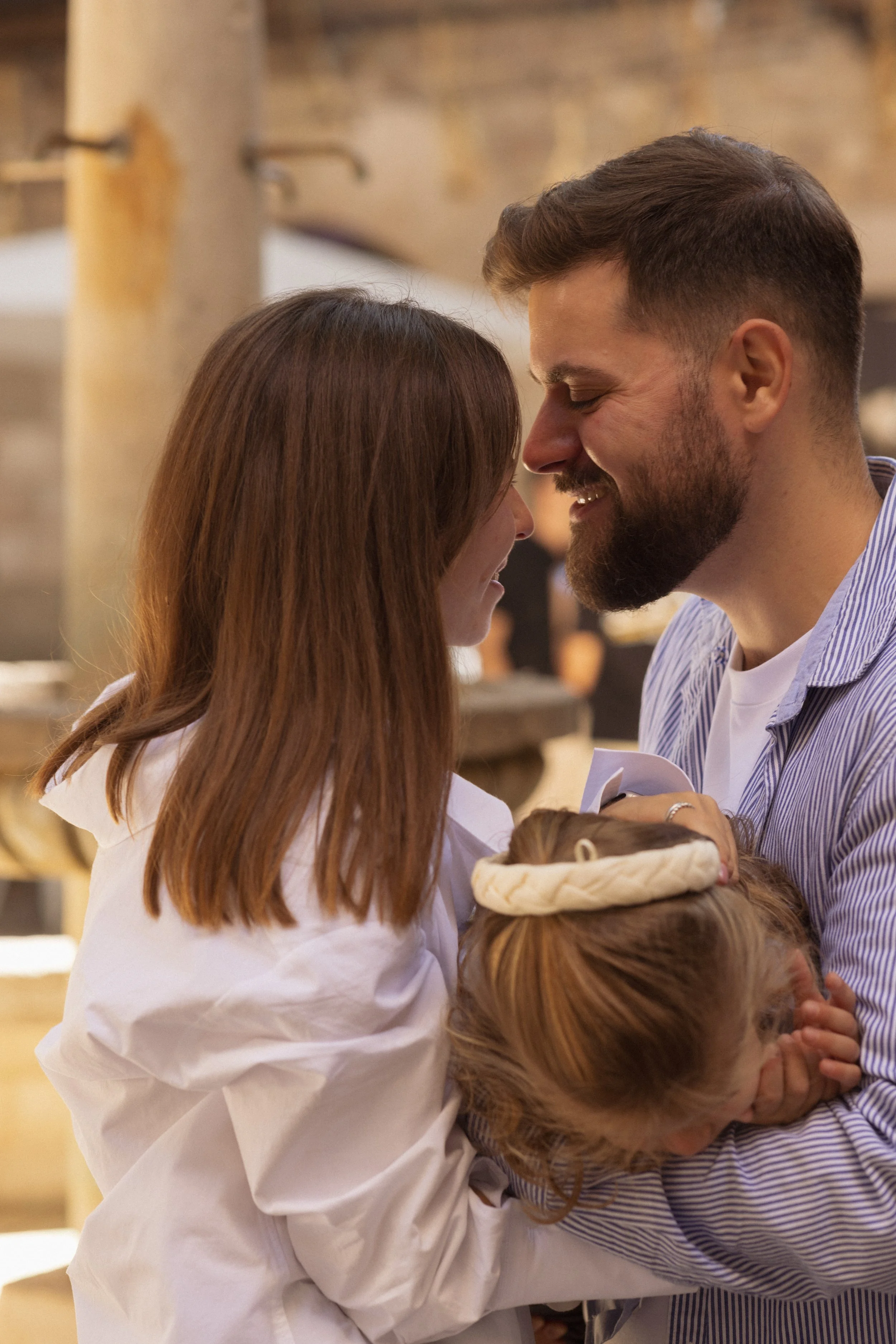 A couple with a young girl sharing a close, happy moment indoors, smiling with foreheads touching.
