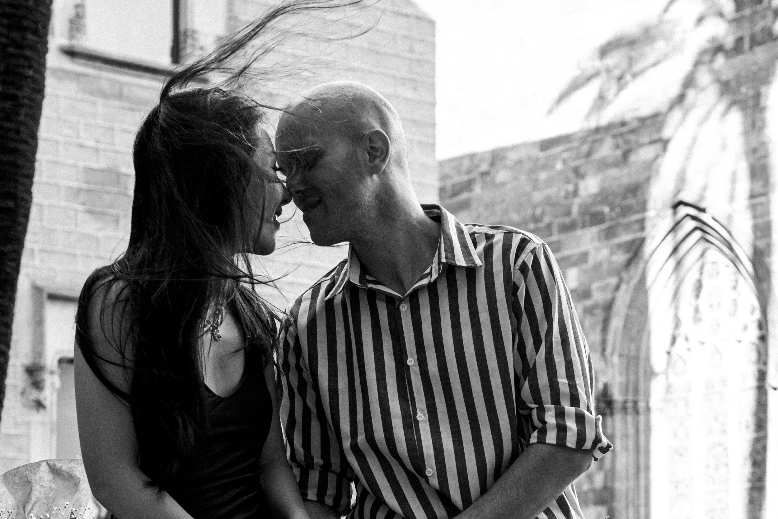 A black-and-white photo of a couple leaning in close, touching foreheads, smiling, outdoors with a brick building and an archway in the background.