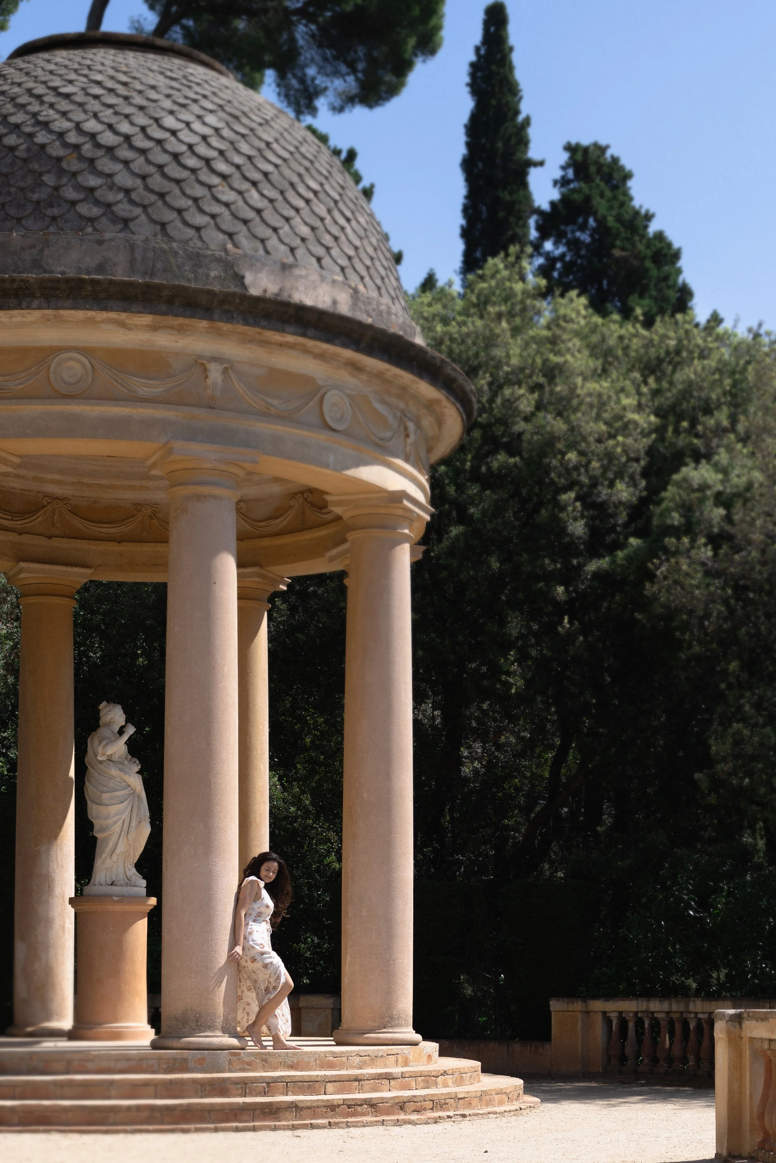 A woman in a floral dress standing on steps under a historic classical pavilion with columns, a statue, and a tiled dome roof, surrounded by trees and clear blue sky.
