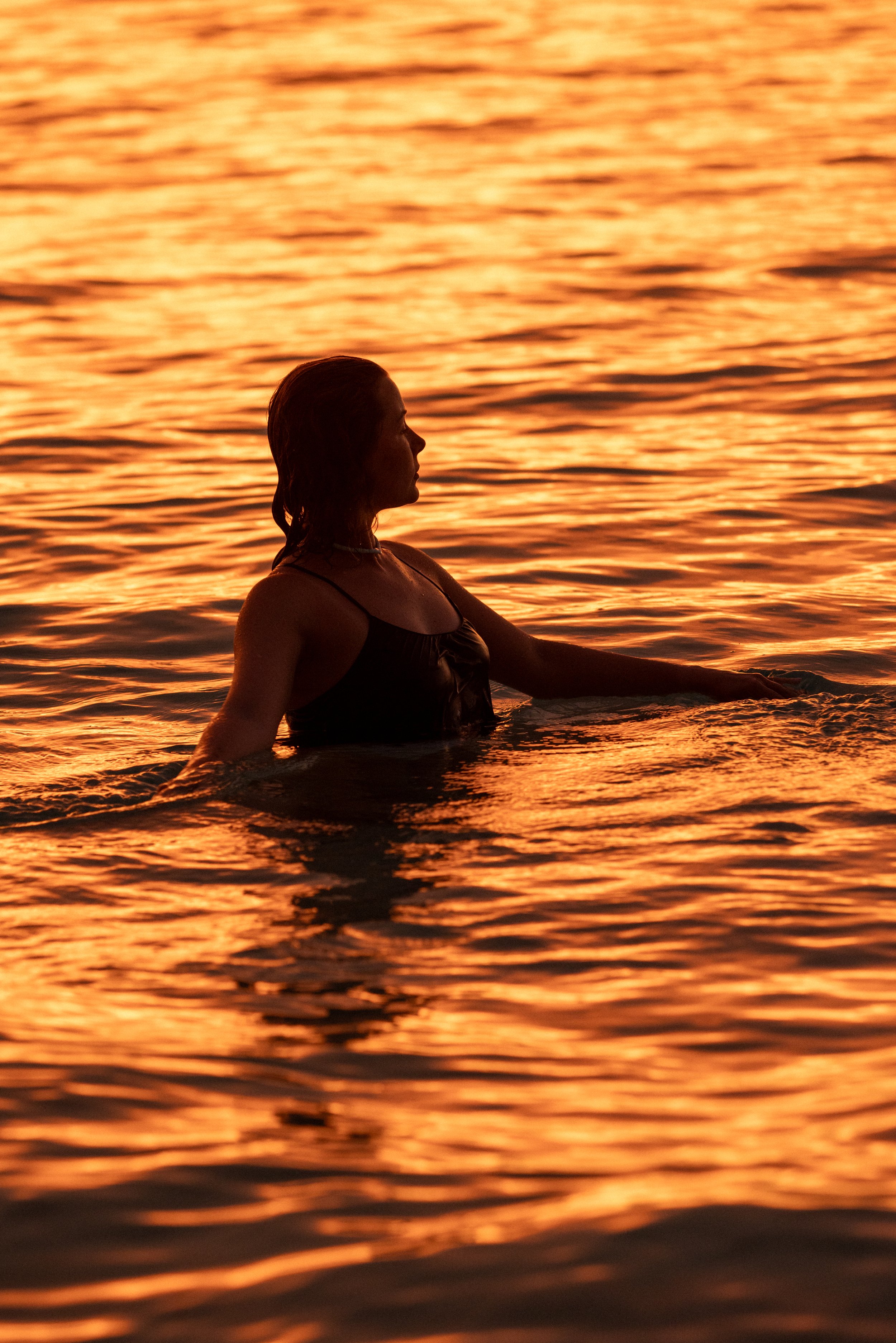 A woman swimming in a body of water during sunset, with the sky and water reflecting a warm orange glow.