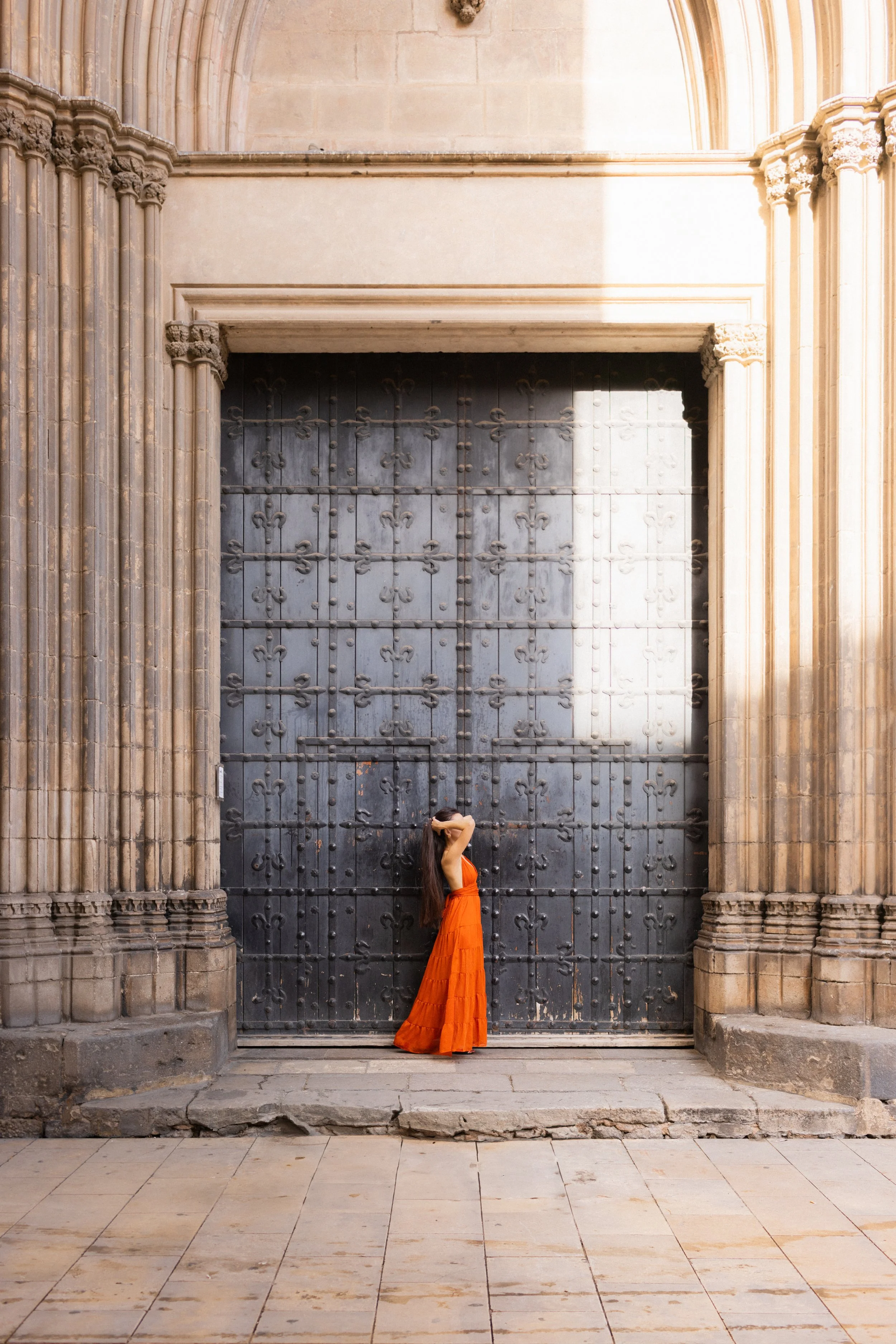 A woman in an orange dress standing in front of a large black historic wooden door with elaborate ironwork, in an old stone building.