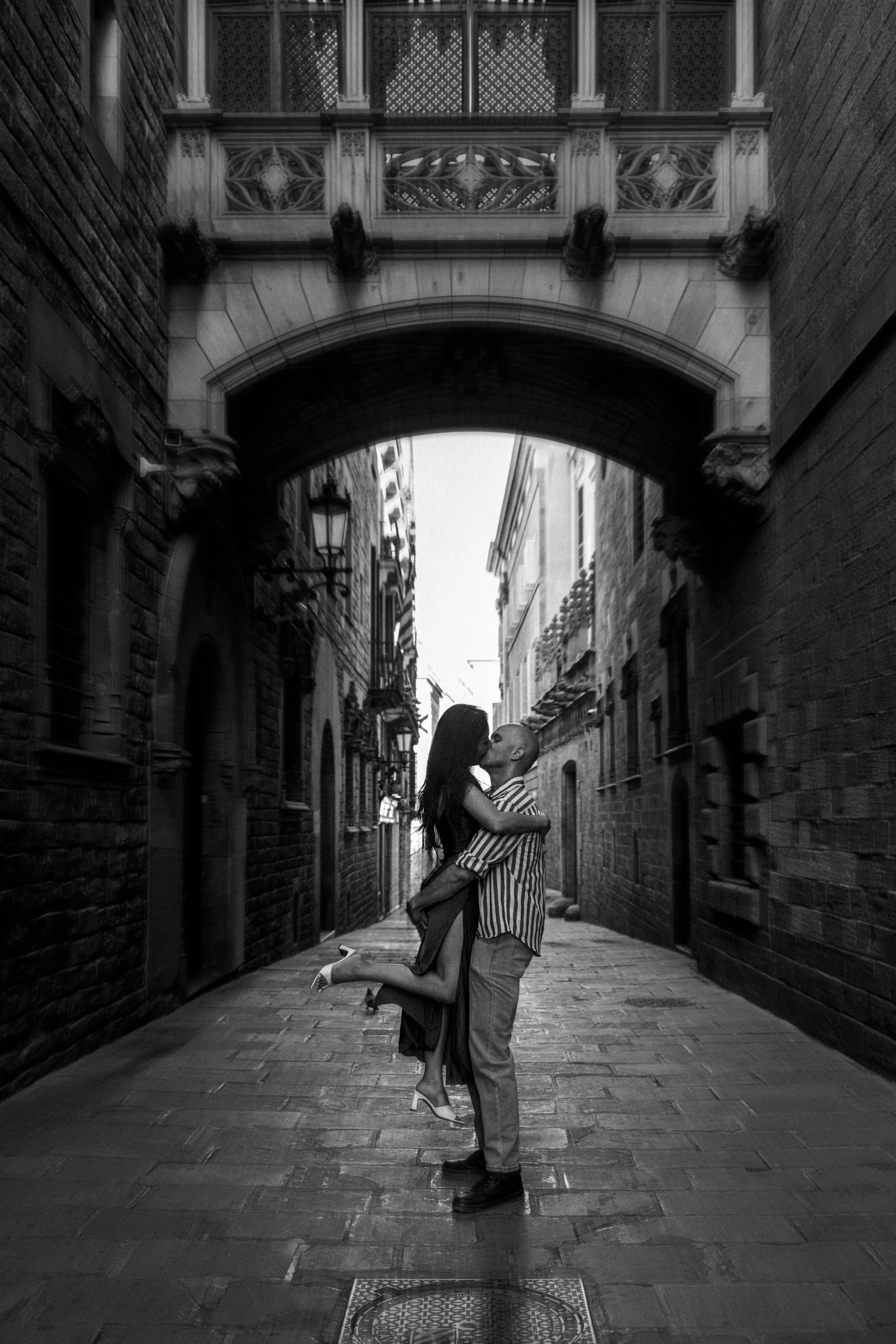 A black and white photo of a couple kissing in a narrow Gothic quarter street with brick walls and an ornate bridge overhead. The woman is lifted off the ground, hugging the man, and wearing high-heeled shoes.