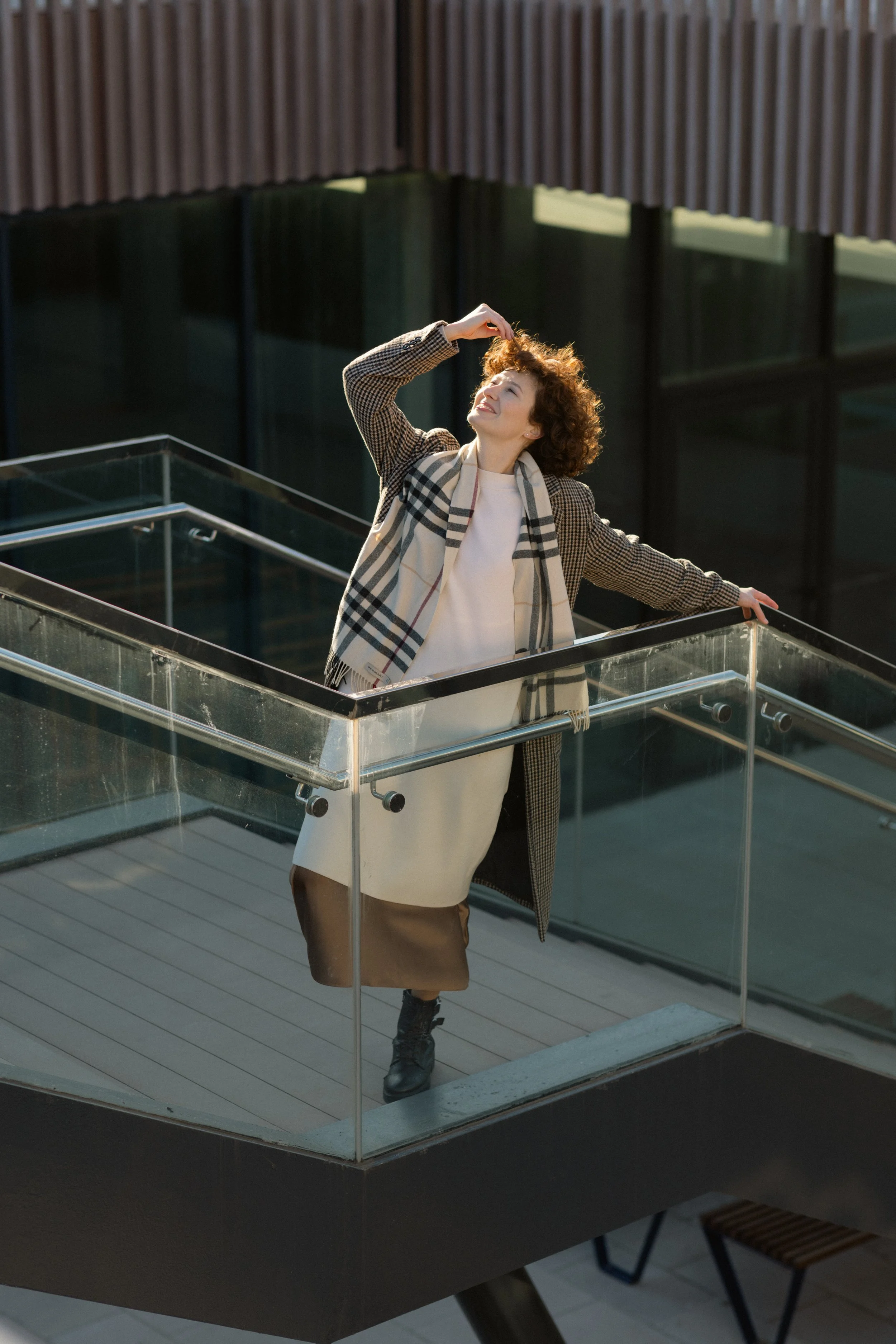 Woman standing on a glass balcony, smiling and touching her curly hair, wearing a beige coat, white dress, and plaid scarf, in an urban setting.