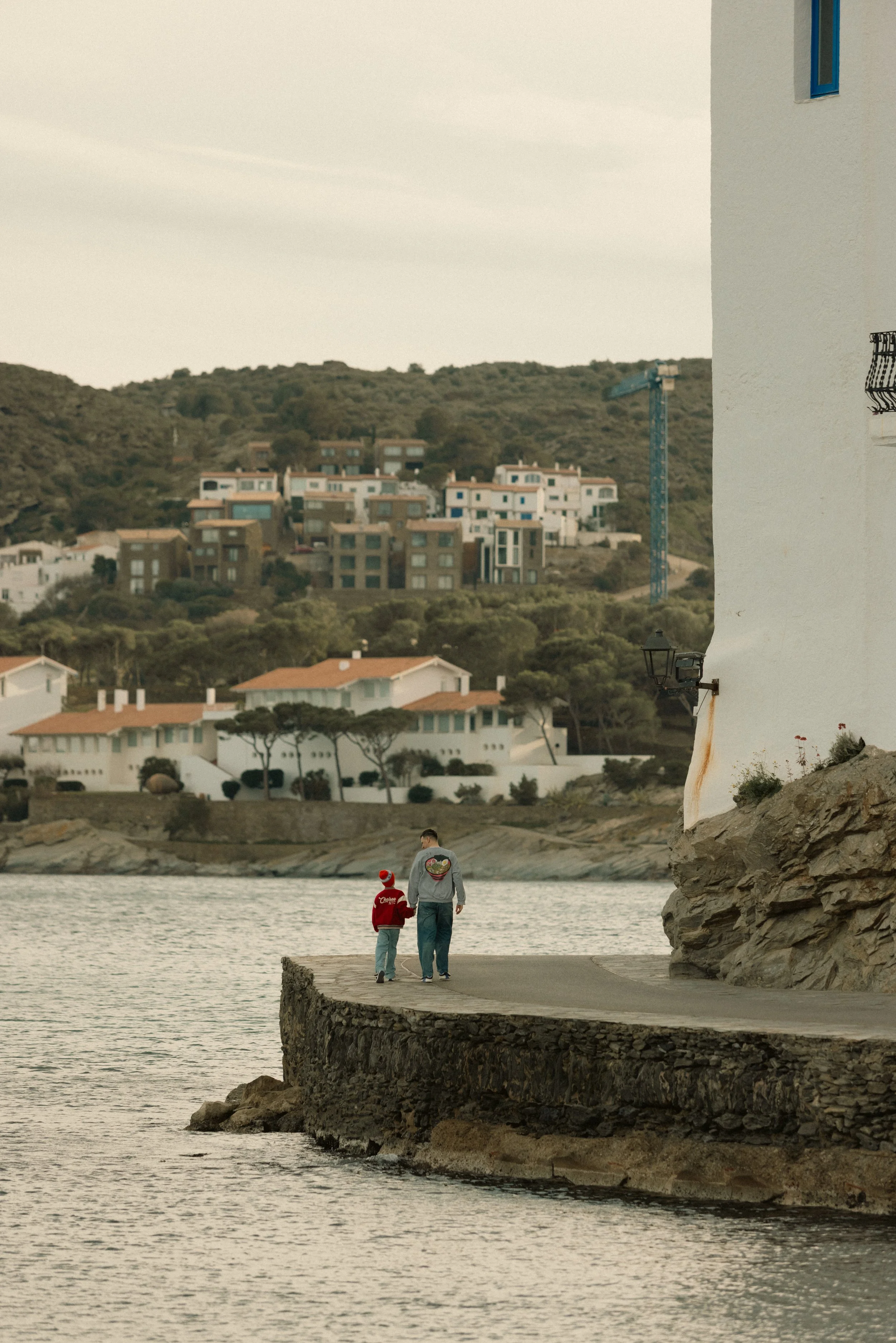 A man and a child walk along a stone path beside the water, with a hillside of houses and buildings in the background.