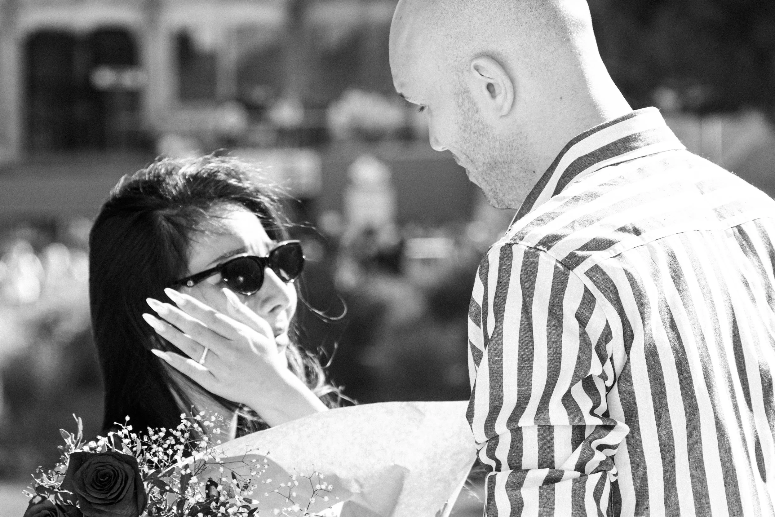 A man presenting a bouquet of flowers to a woman wearing sunglasses, outdoors on a sunny day.