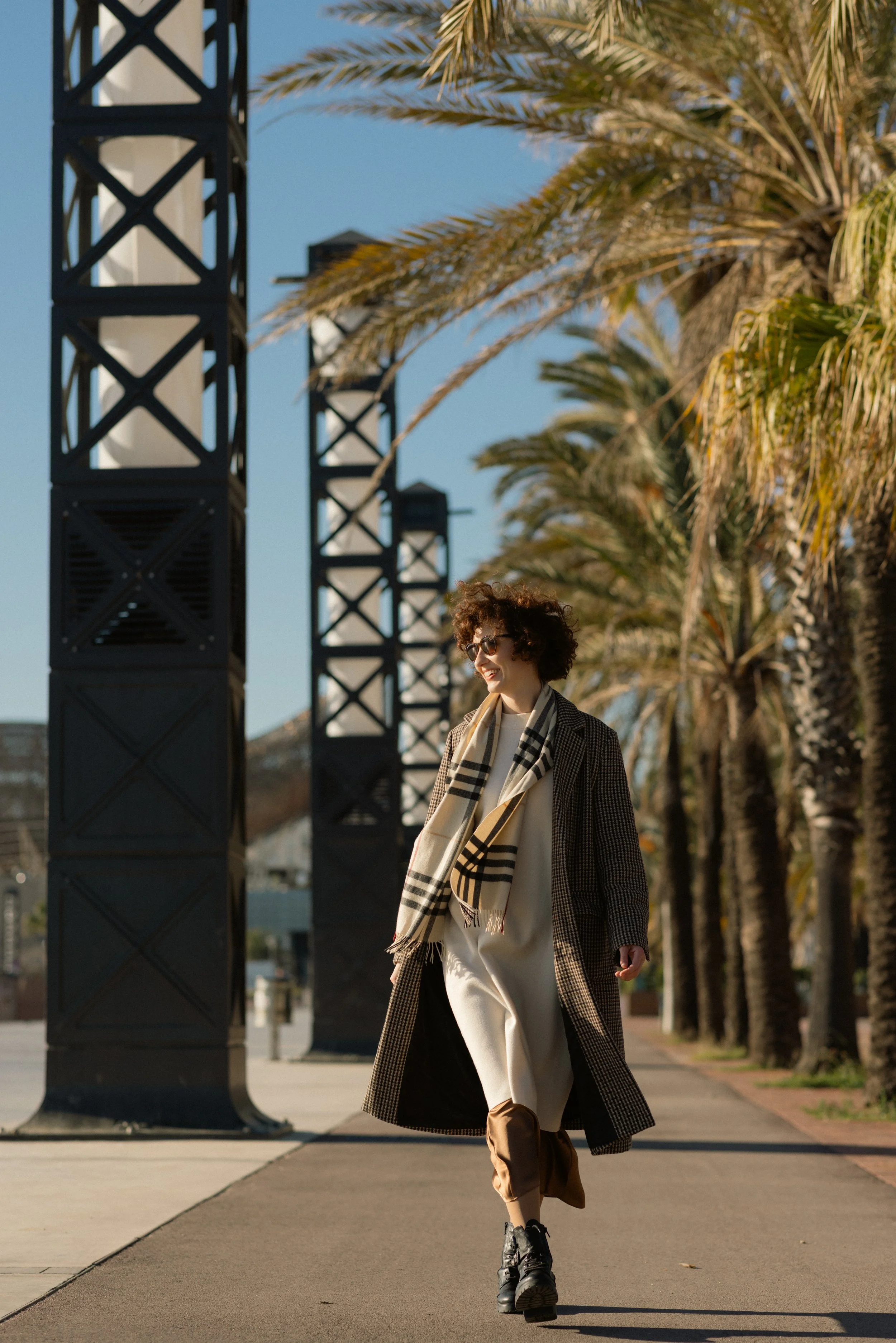 A woman walking along a sunny city street lined with palm trees, wearing sunglasses, a long coat, and a scarf.
