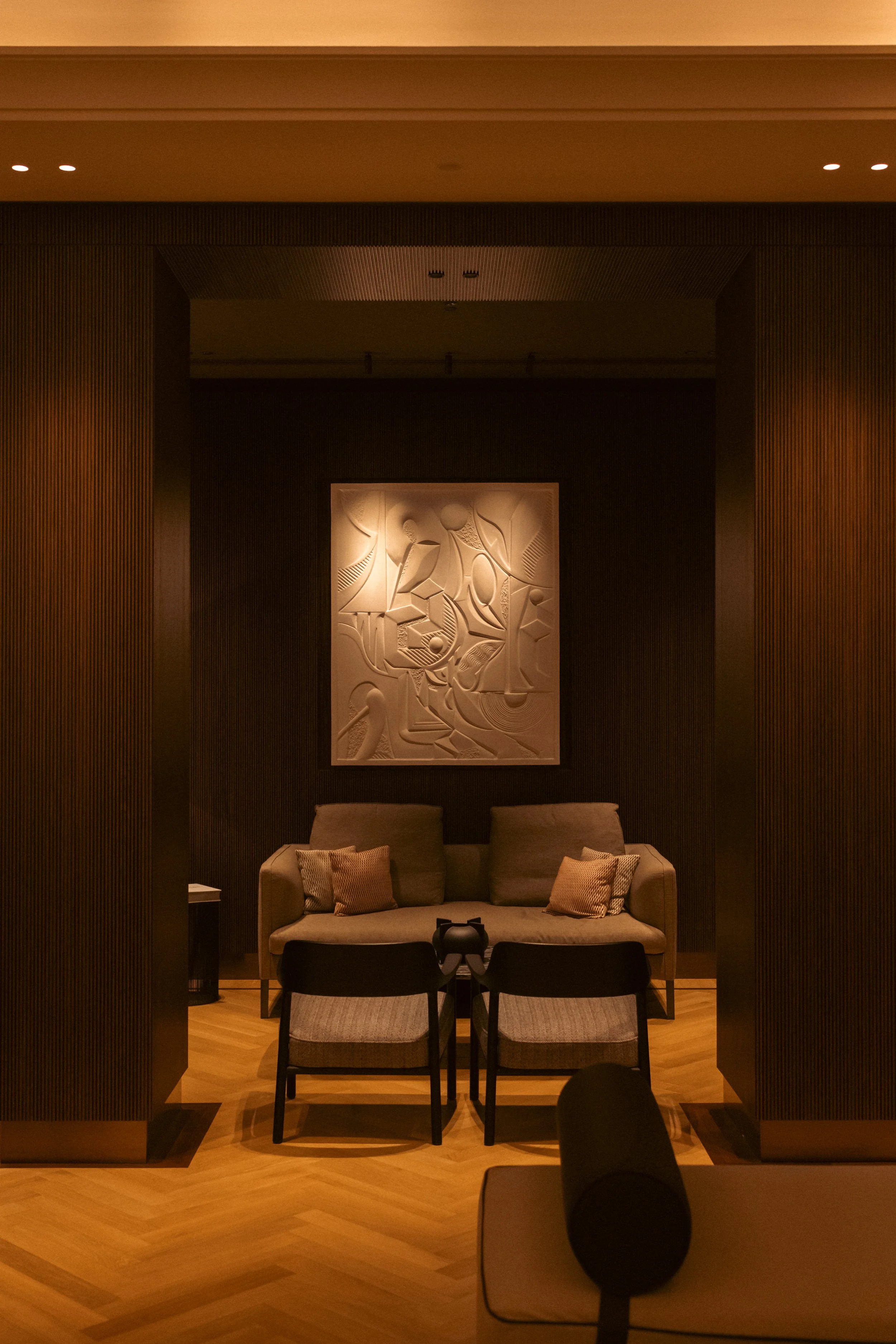 Living room area with two chairs, a sofa with pillows, two decorative chairs, a framed abstract artwork, wooden panel walls, and a wooden floor.