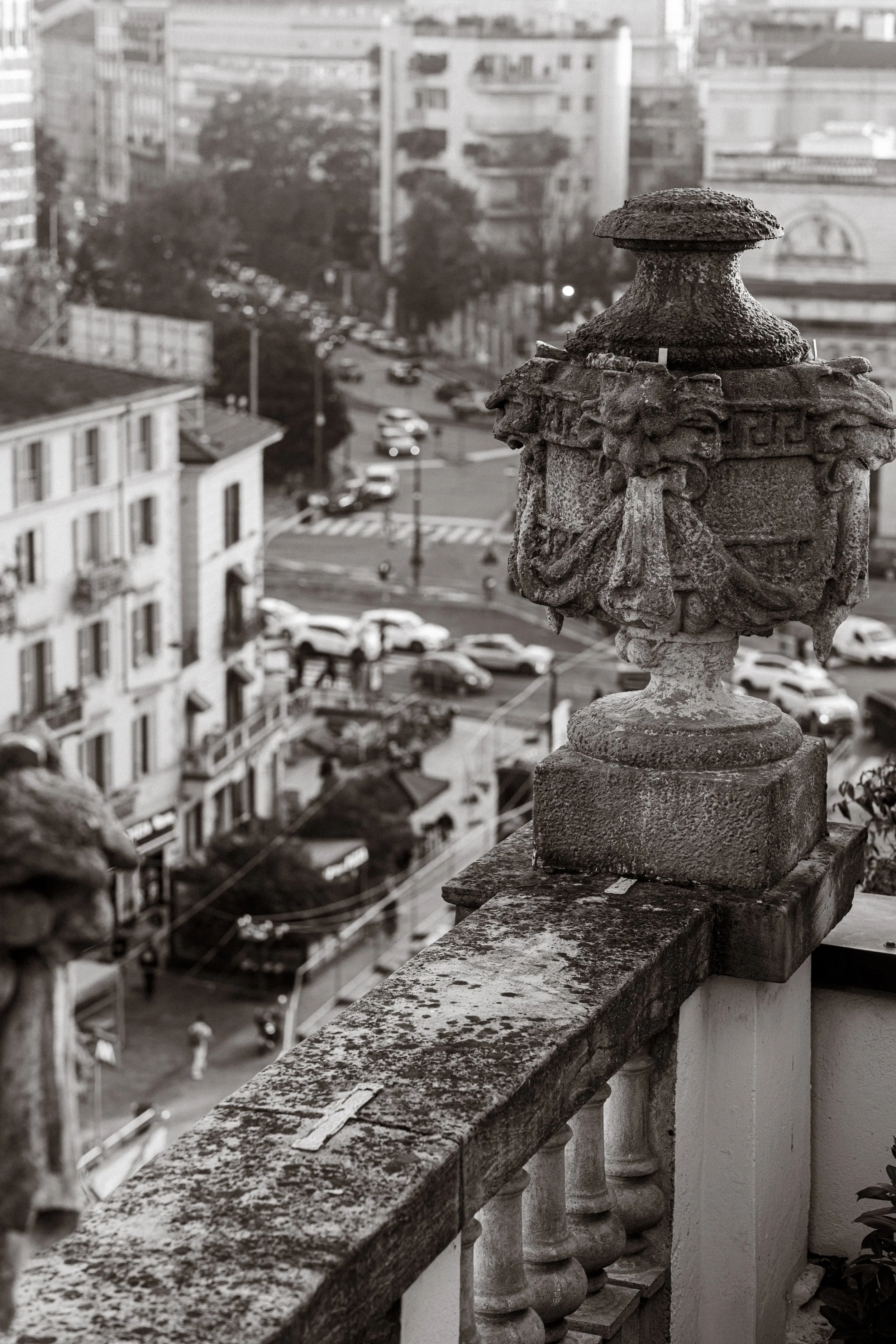 Close-up of an ornate stone balustrade with a decorative stone urn on top, overlooking a city street with cars and buildings in the background in black and white.