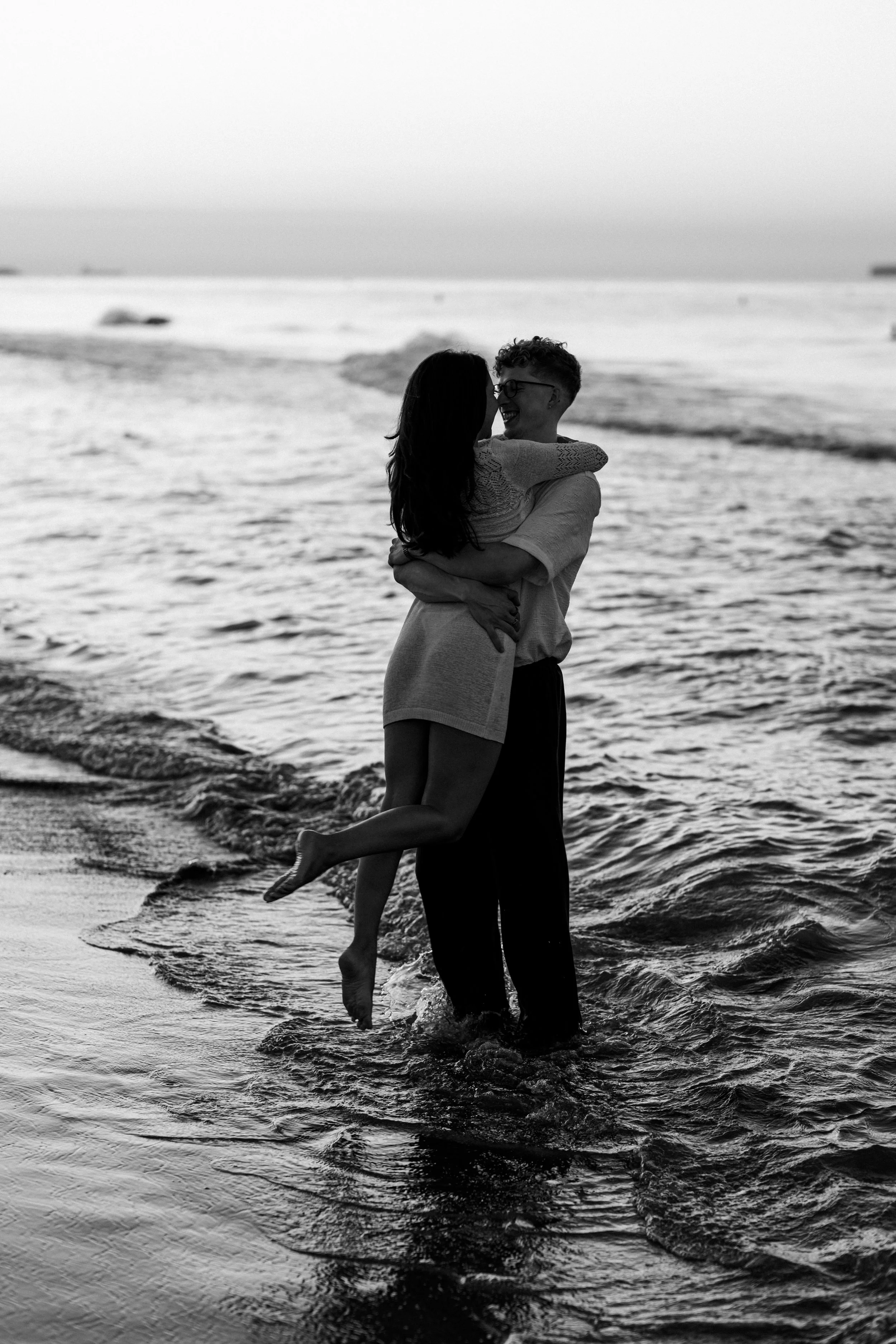 A black and white photo of a couple hugging in the ocean water at the beach, with the woman being lifted off the ground and both laughing.