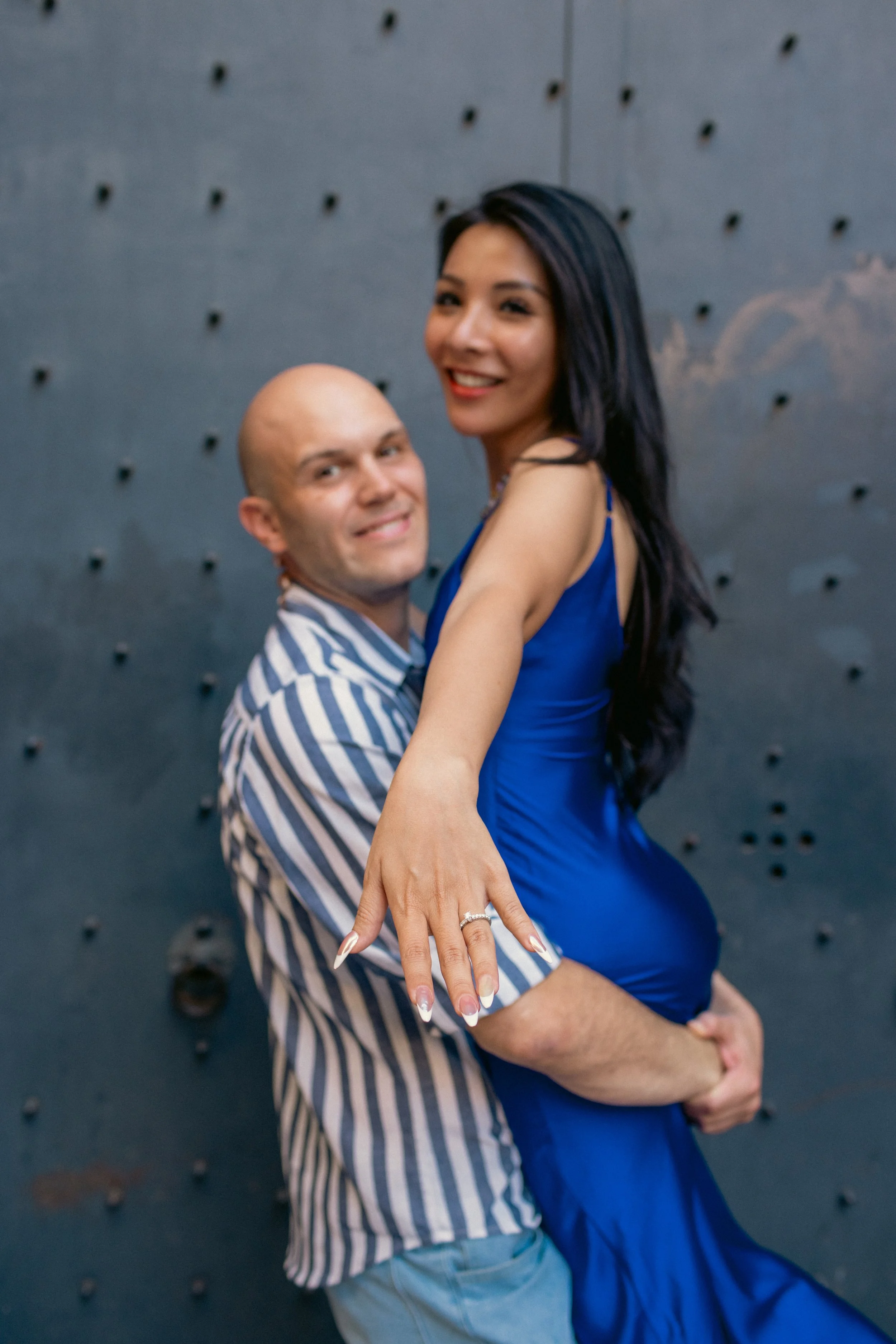 A joyful couple poses in front of a textured metal wall, with the woman wearing a fitted blue dress showing an engagement ring, and the man lifting her while smiling at the camera.