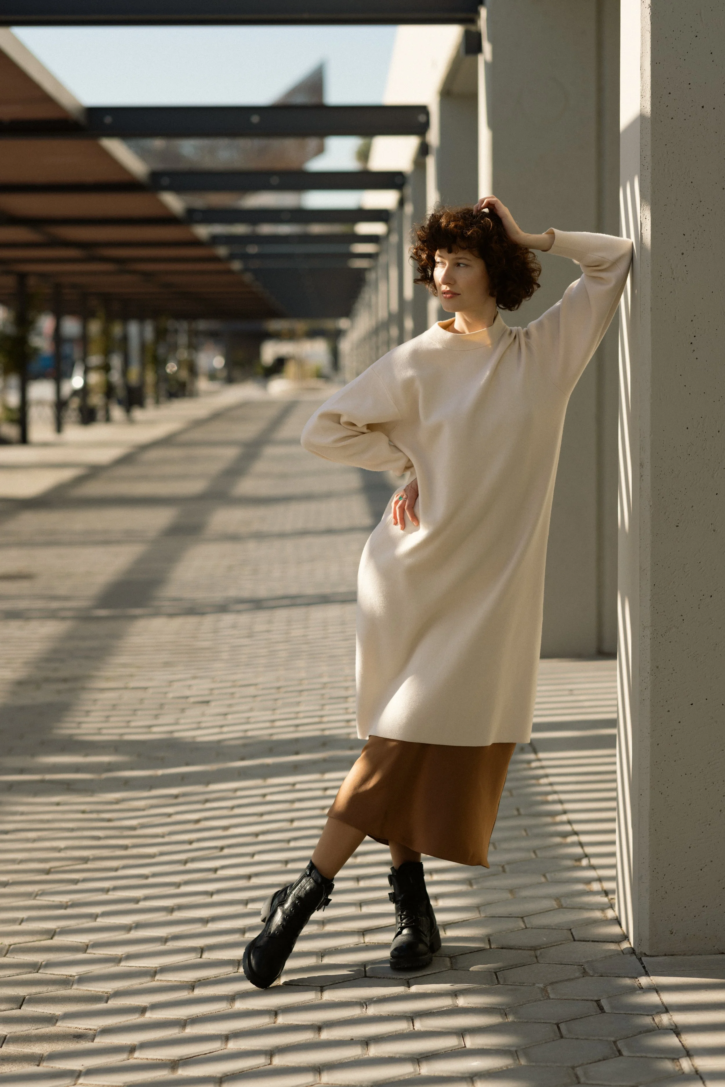 A woman in a beige oversized sweater, brown skirt, black combat boots, standing outdoors against a cement wall, with shadows cast on the sidewalk during daylight.
