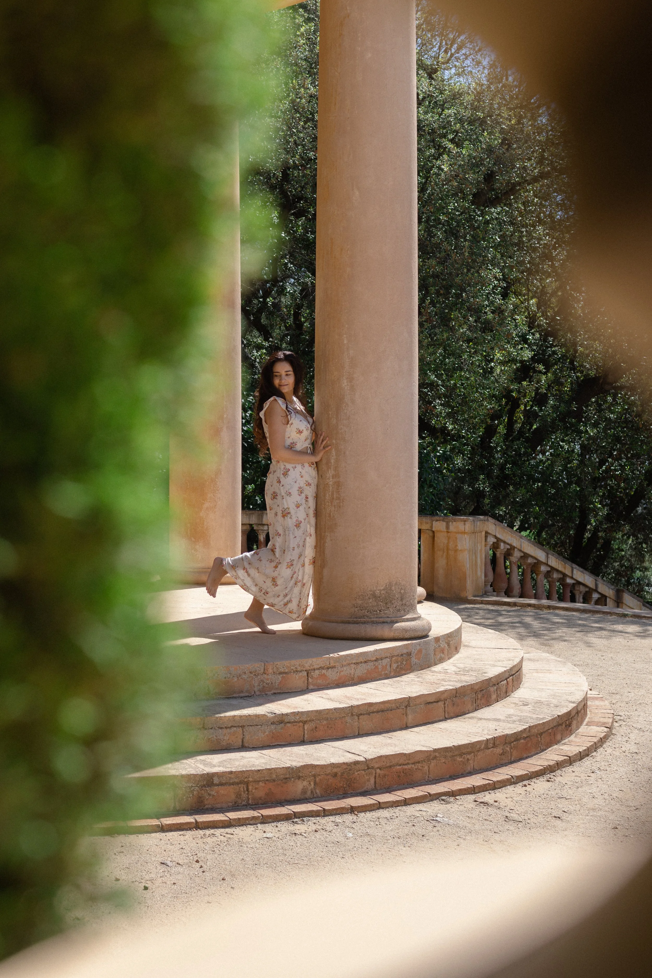 A woman in a floral dress leaning against a stone column on a round staircase in an outdoor setting, surrounded by greenery.
