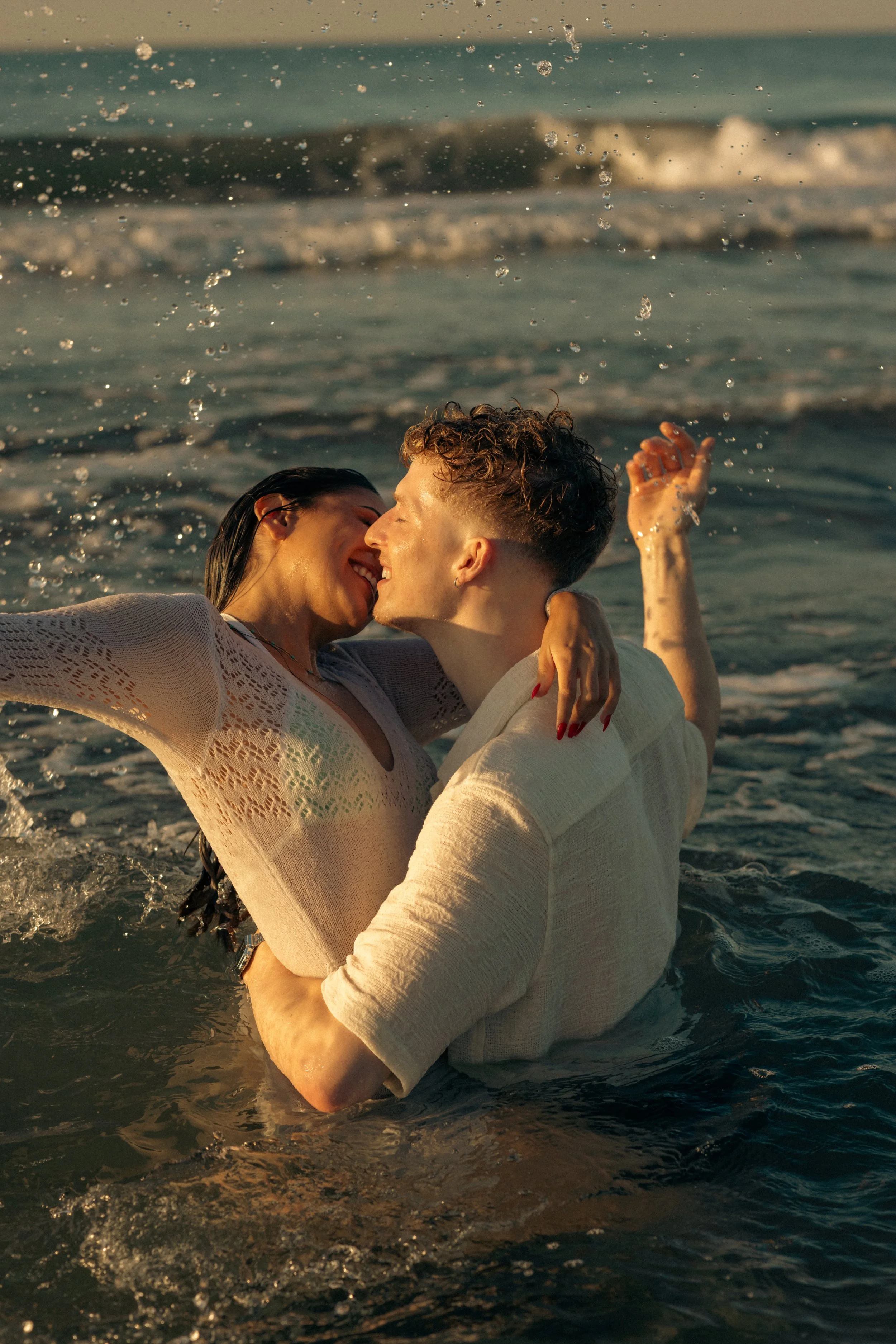 A couple kissing in the ocean during sunset, embracing in shallow water with waves around them.